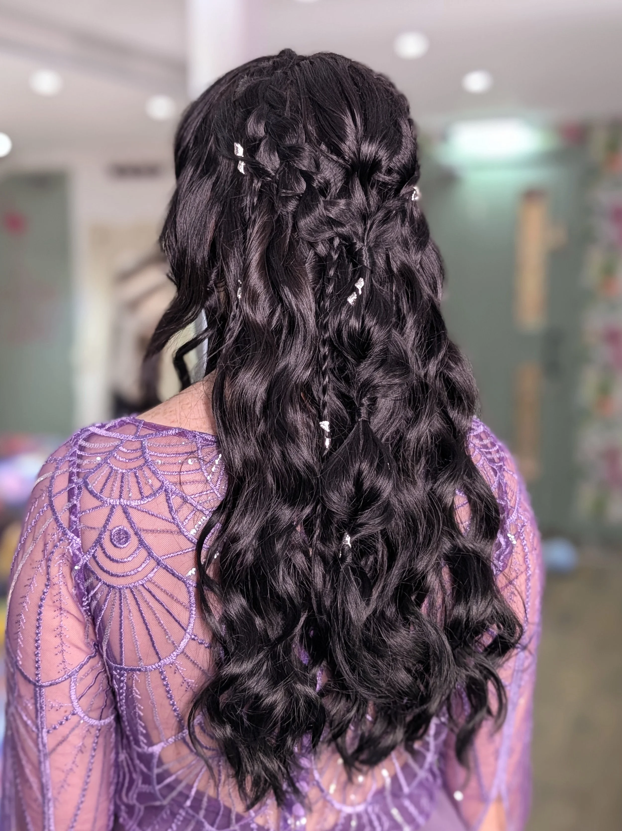 Back view of a woman with long, dark, wavy hair with small decorative clips, wearing a purple dress with intricate embroidery.