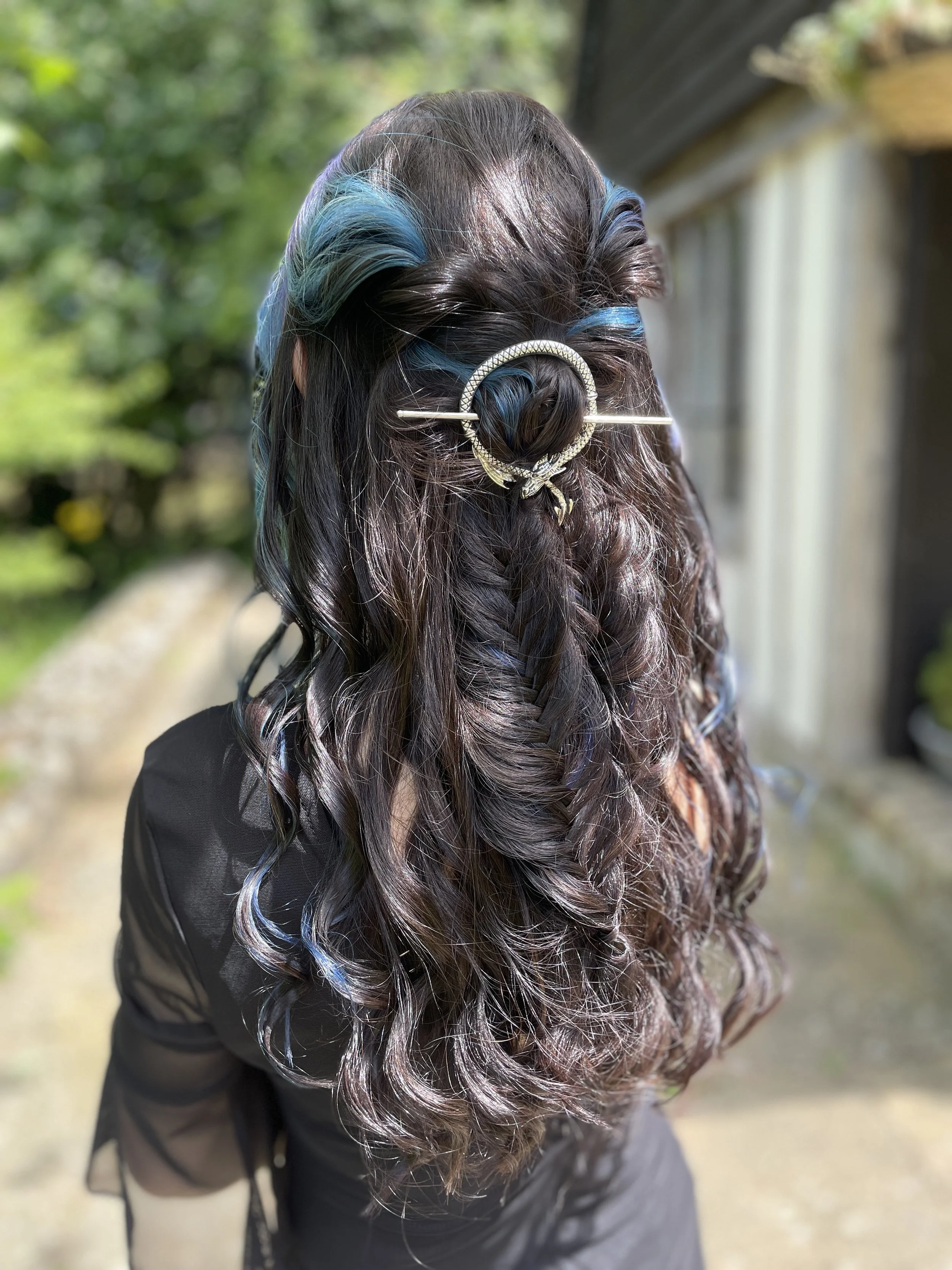 Back view of a woman with long, dark, curly hair with blue highlights, styled with a decorative hair clip in the shape of a circle with a bow, outdoors in sunlight.
