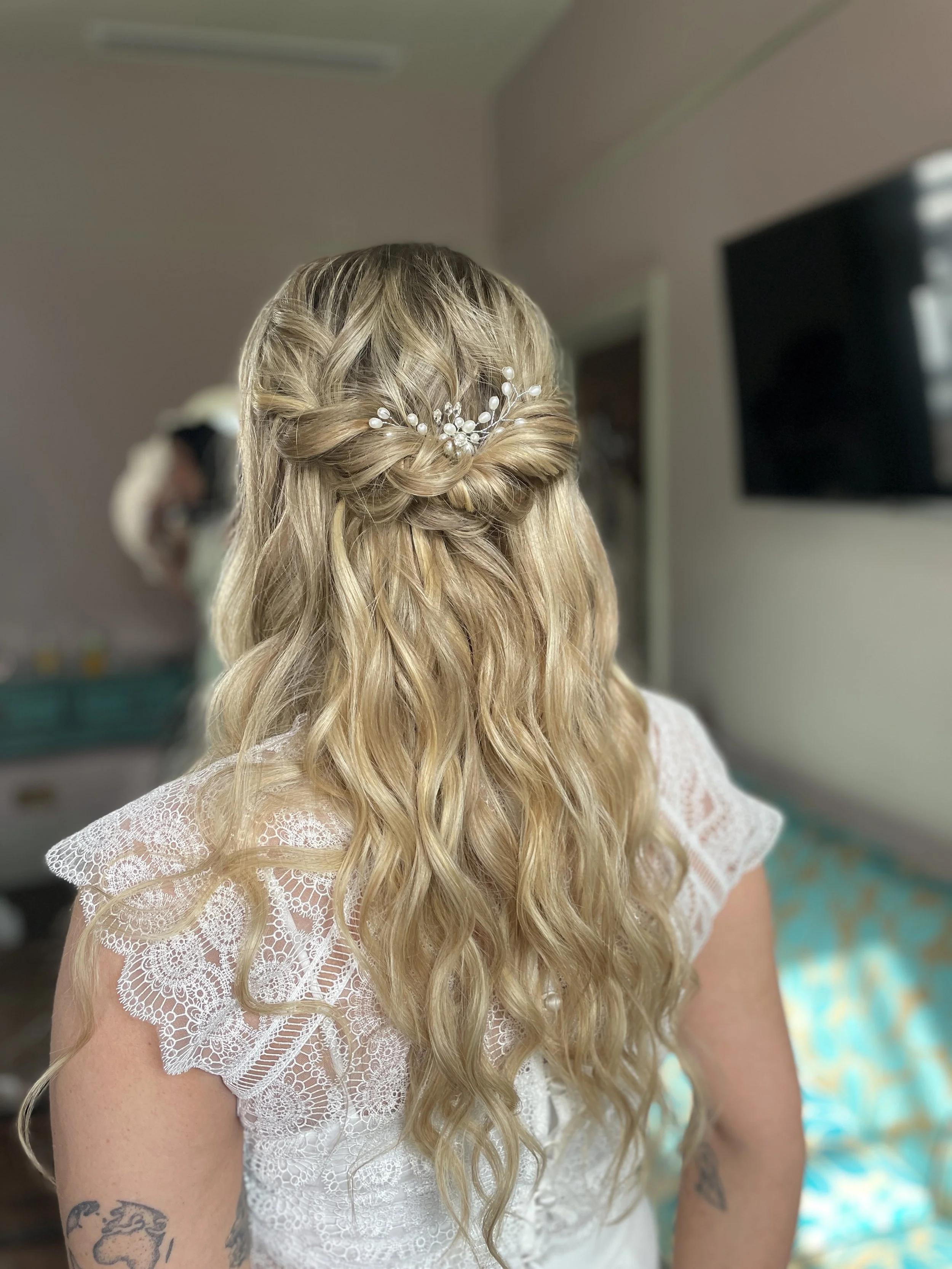 Back view of a woman with long, wavy blonde hair styled with a small twisted braid across the crown, decorated with a pearl and bead hair accessory, wearing a white lace dress with short sleeves.