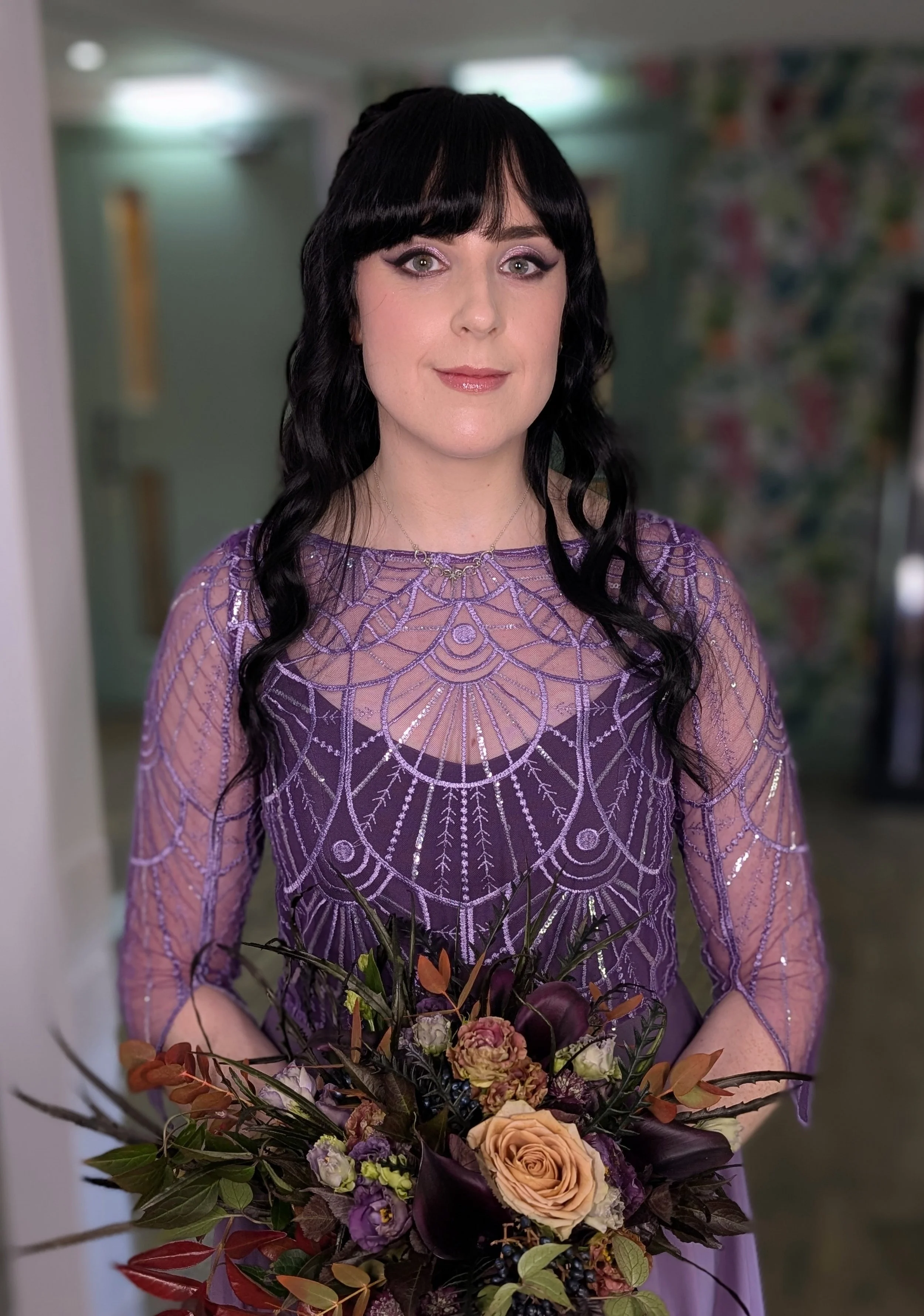 A woman with black wavy hair and fair skin, wearing a purple dress with intricate silver embroidery, holds a bouquet of mixed flowers, including roses, in various colors and dark leaves.