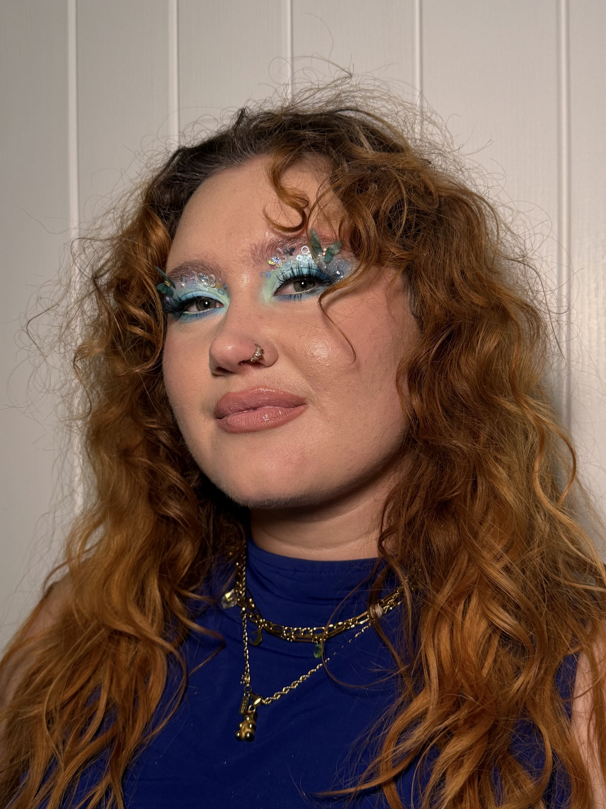 Close-up of a woman with curly red hair wearing blue clothing and layered necklaces, with elaborate colorful makeup and rhinestones around her eyes.