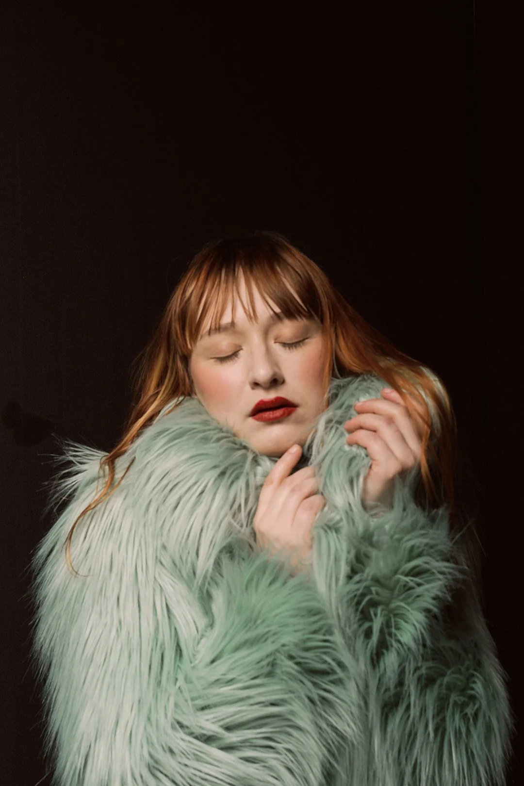 A woman with reddish hair and red lipstick, wearing a fluffy light green coat, on a dark background with her eyes closed and her hands near her face.
