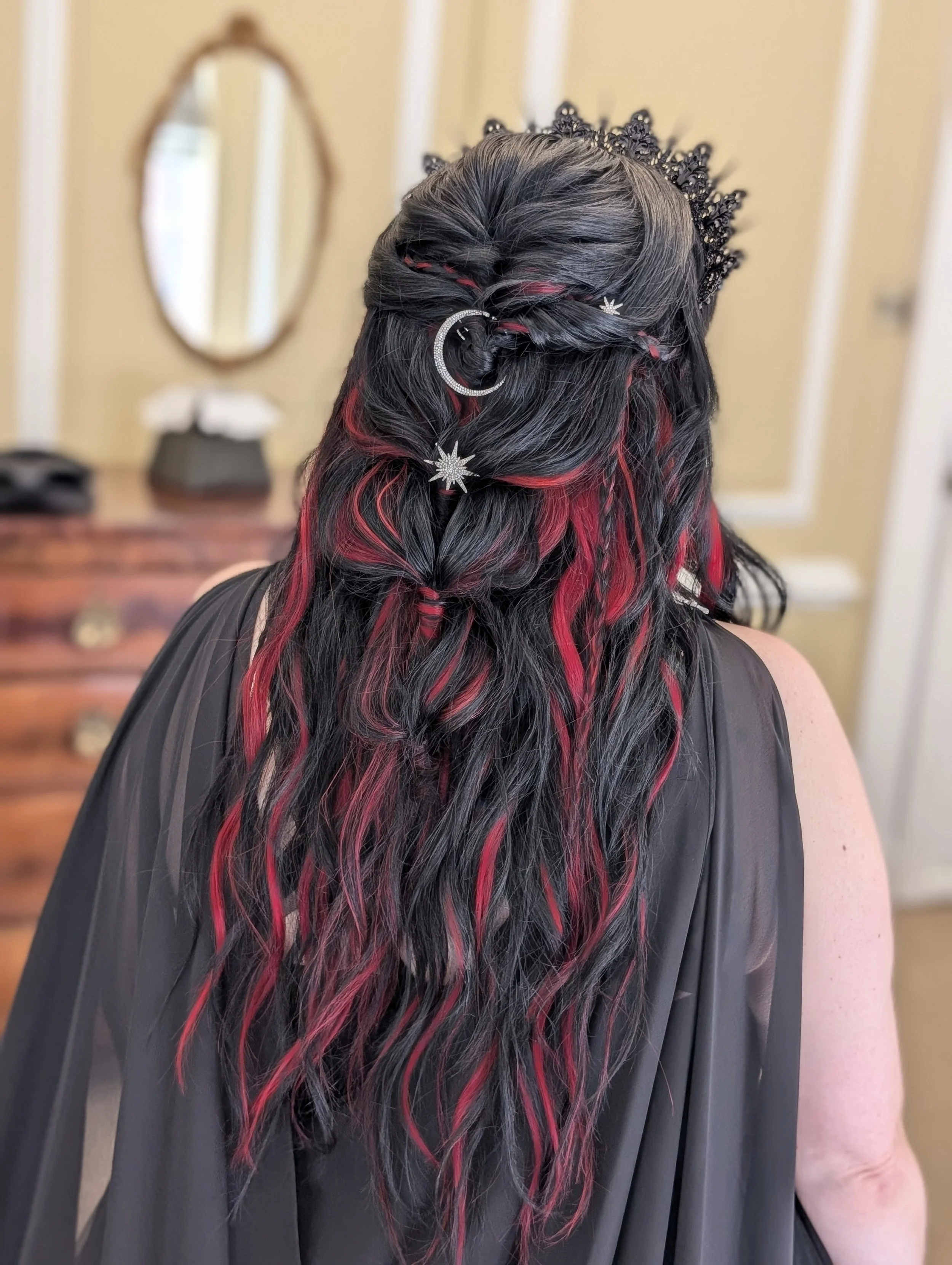 Back view of a woman with long black hair with red streaks, styled with decorative black accessories and star-shaped jewelry, wearing a black draped garment.
