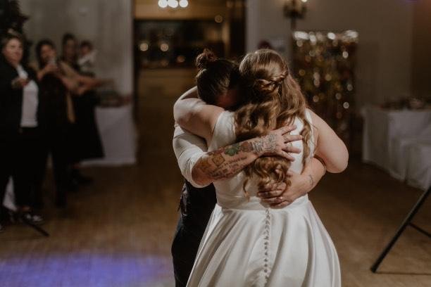 Two women hugging during a wedding reception in a decorated indoor venue, with guests in the background.