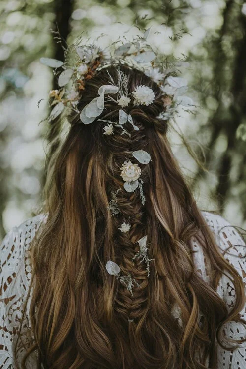 Woman with long, wavy hair styled in a braid, wearing a floral crown with white flowers and leaves, outdoors with blurred trees in the background.
