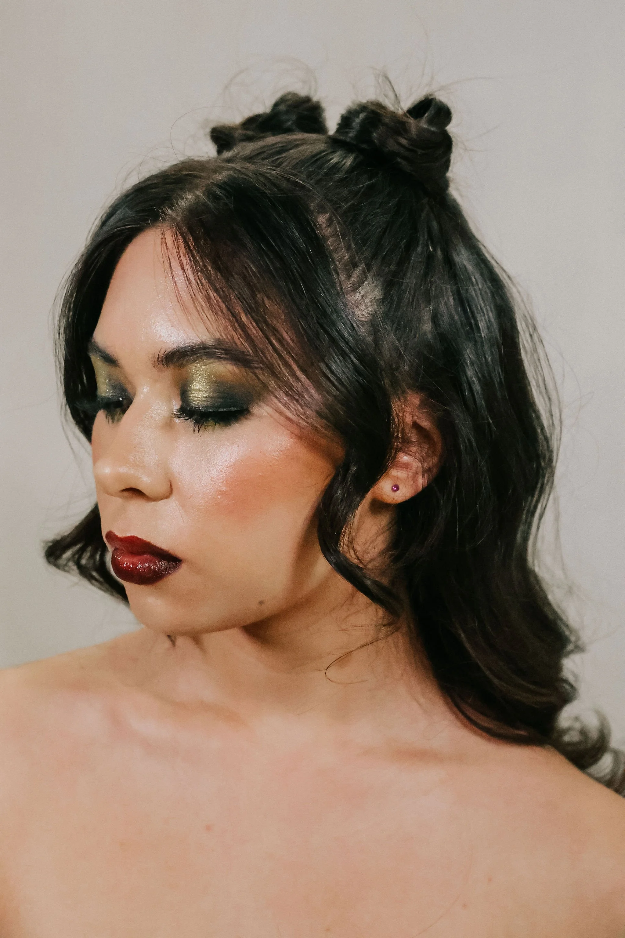 A woman with dark, wavy hair styled in two small buns on top, wearing dark red lipstick, eye makeup with gold and black eyeshadow, and stud earrings, with bare shoulders against a plain background.
