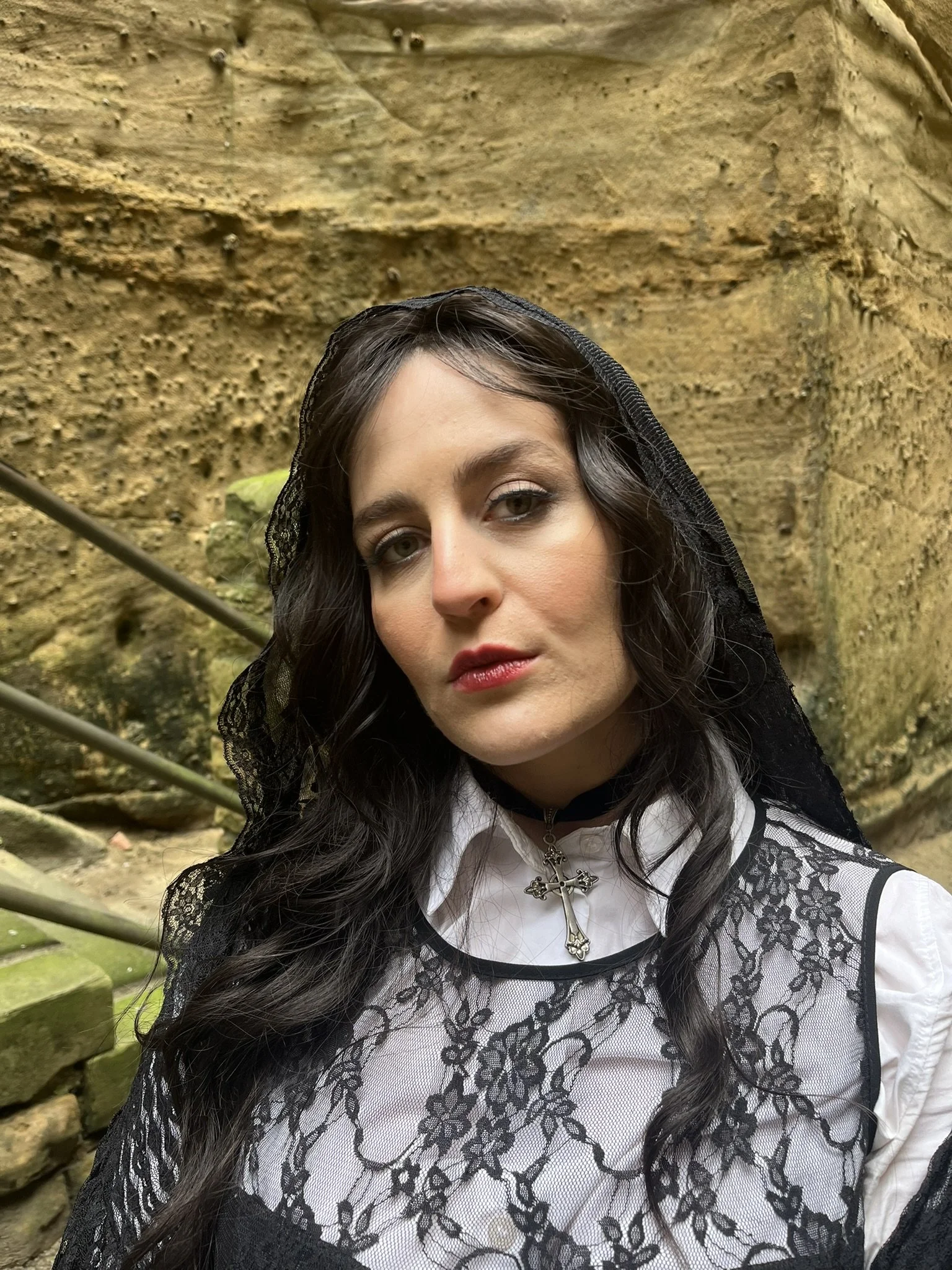 A woman with long dark hair wearing a white blouse with black lace overlay and a black veil, standing outdoors in front of a rocky, moss-covered wall.