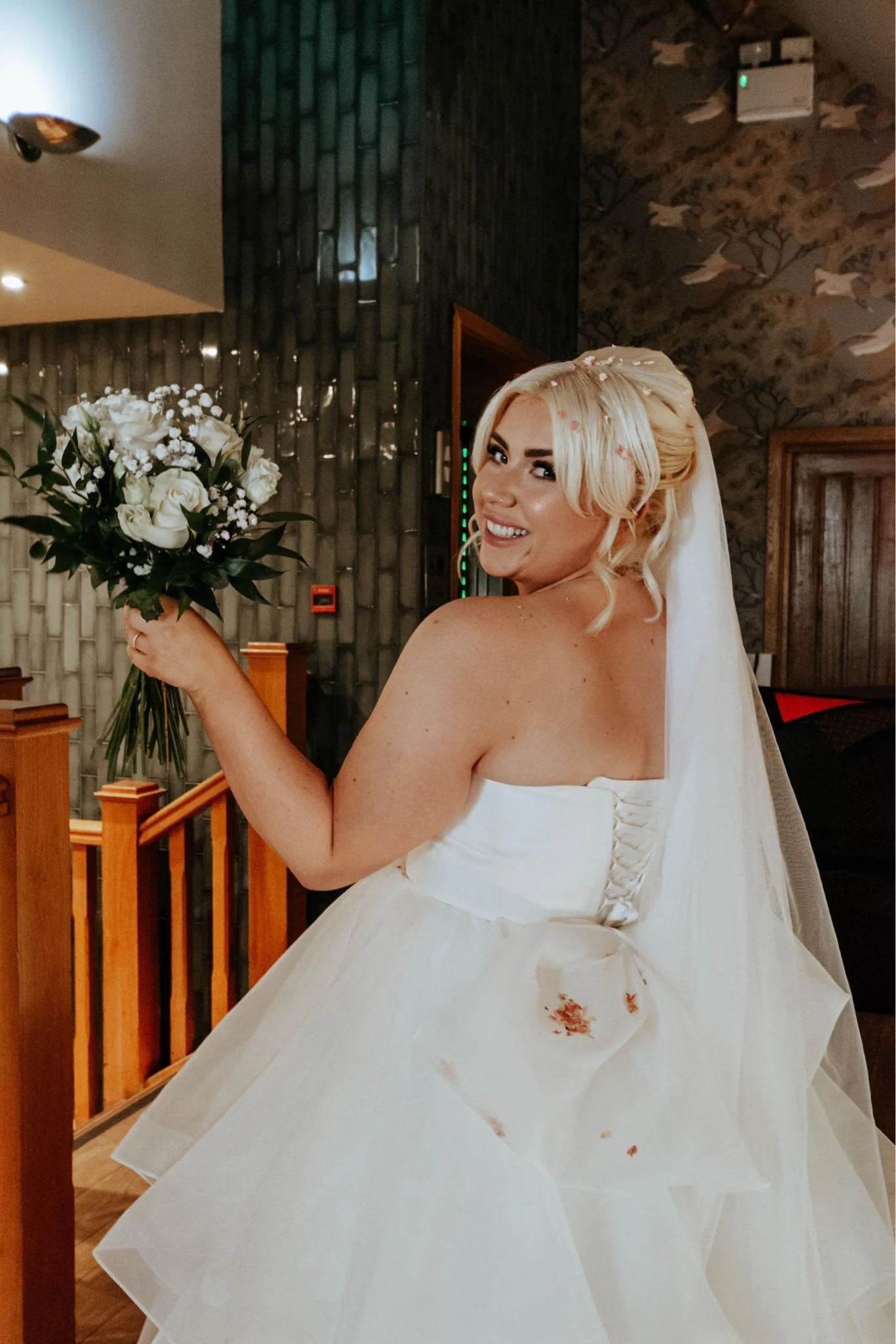 A smiling bride with blonde hair holding a bouquet of white roses and baby's breath, wearing a strapless white wedding dress with a veil, indoors with a textured wall and wooden accents in the background.