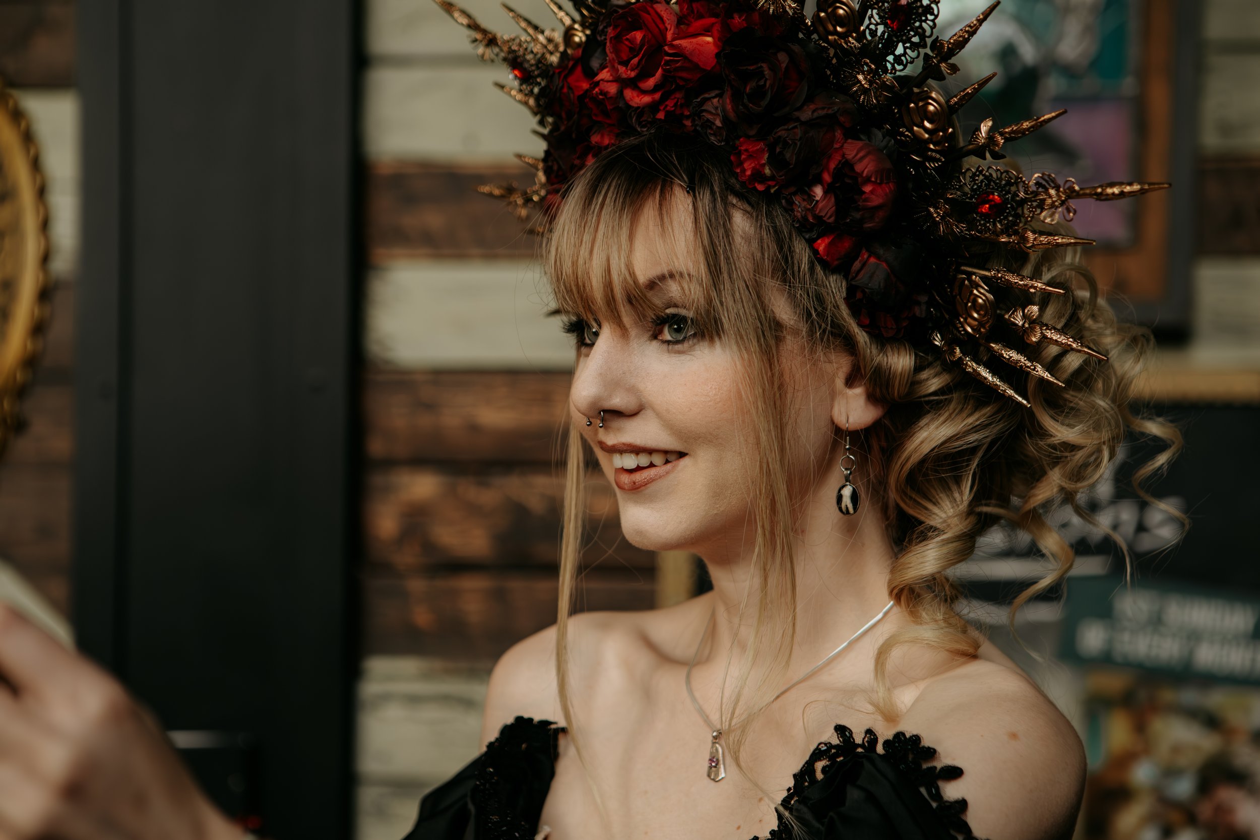 A woman with curly blonde hair wearing a gothic-style headpiece with red and black flowers and spikes, smiling, with piercings and jewelry, against a background of wooden panels.