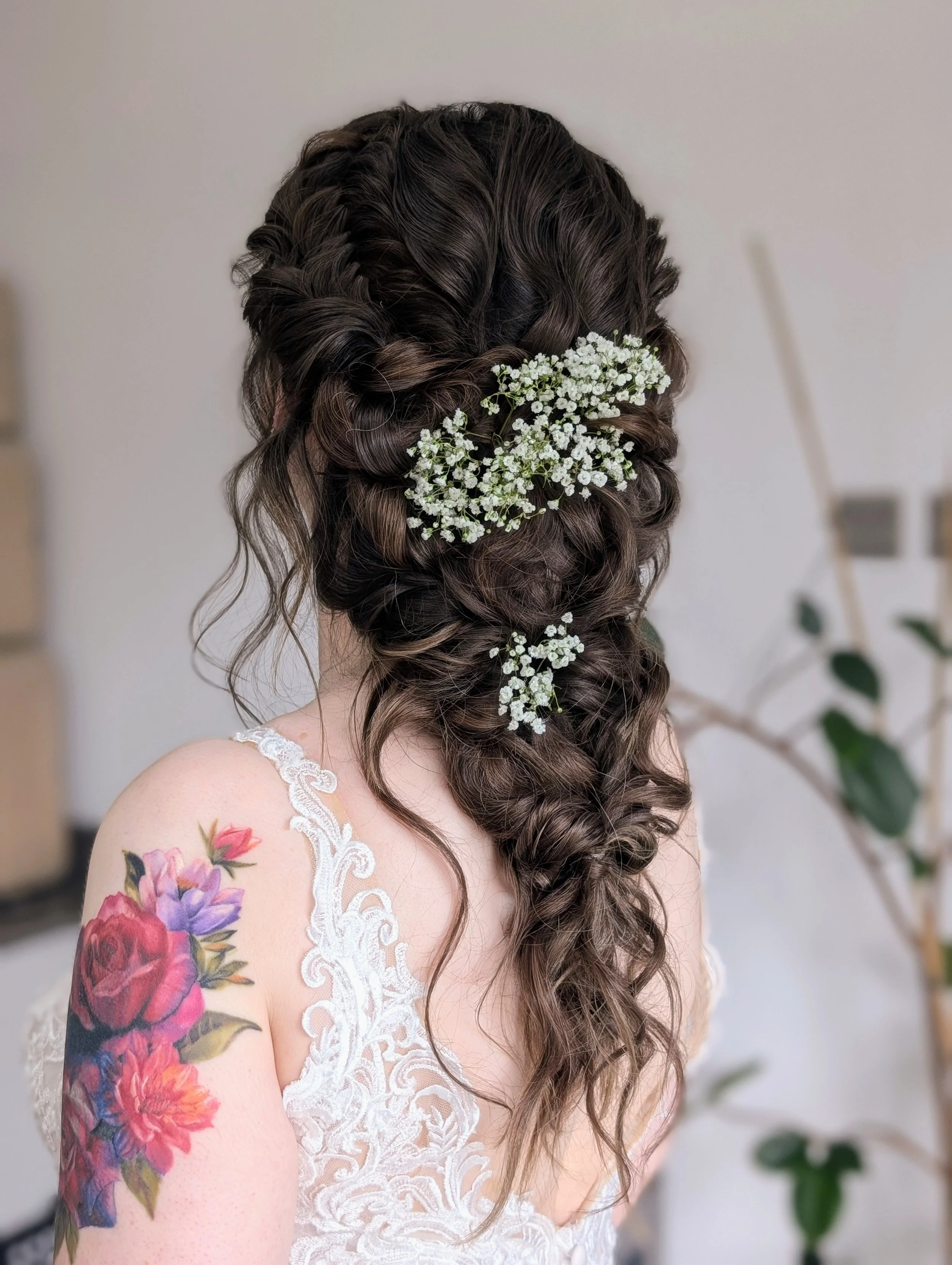 A woman with long, curly brown hair decorated with white baby's breath flowers, wearing a lace wedding dress, showing a colorful floral tattoo on her shoulder.