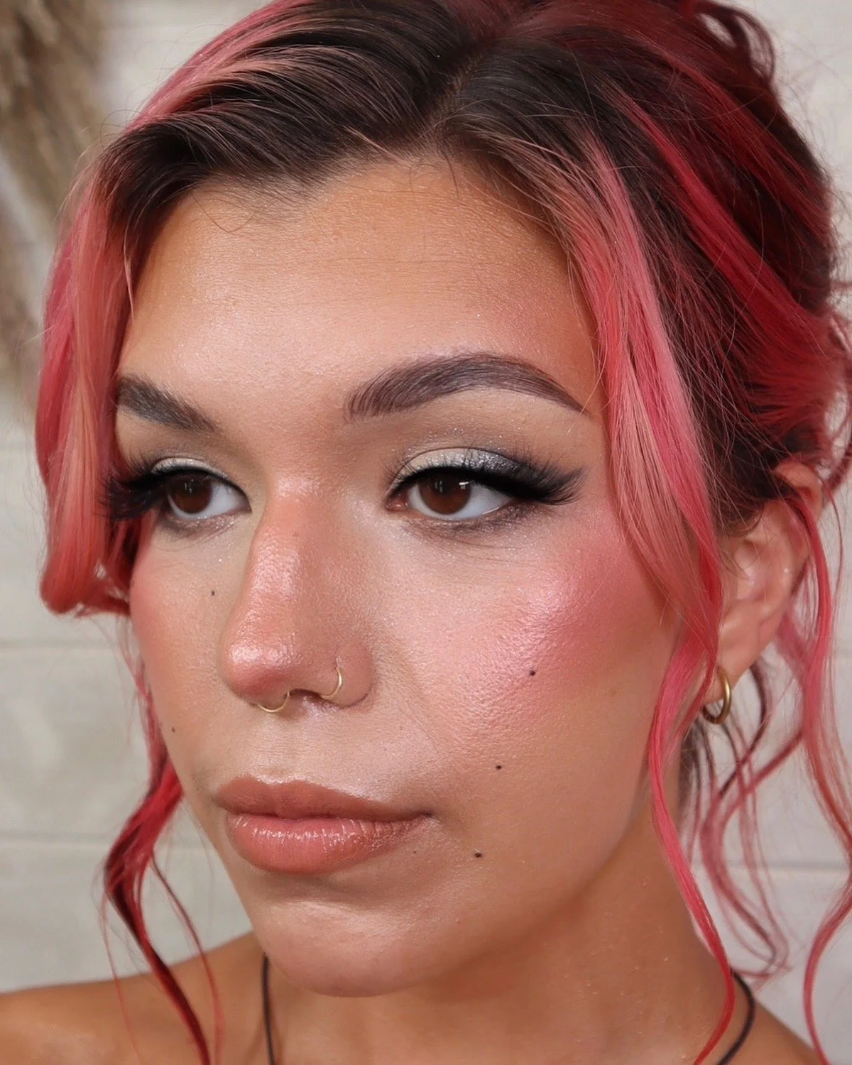 Close-up of a woman's face with pink hair, brown eyes, and makeup including silver eyeshadow, black eyeliner, and nude lipstick. She has a gold nose ring and earrings.