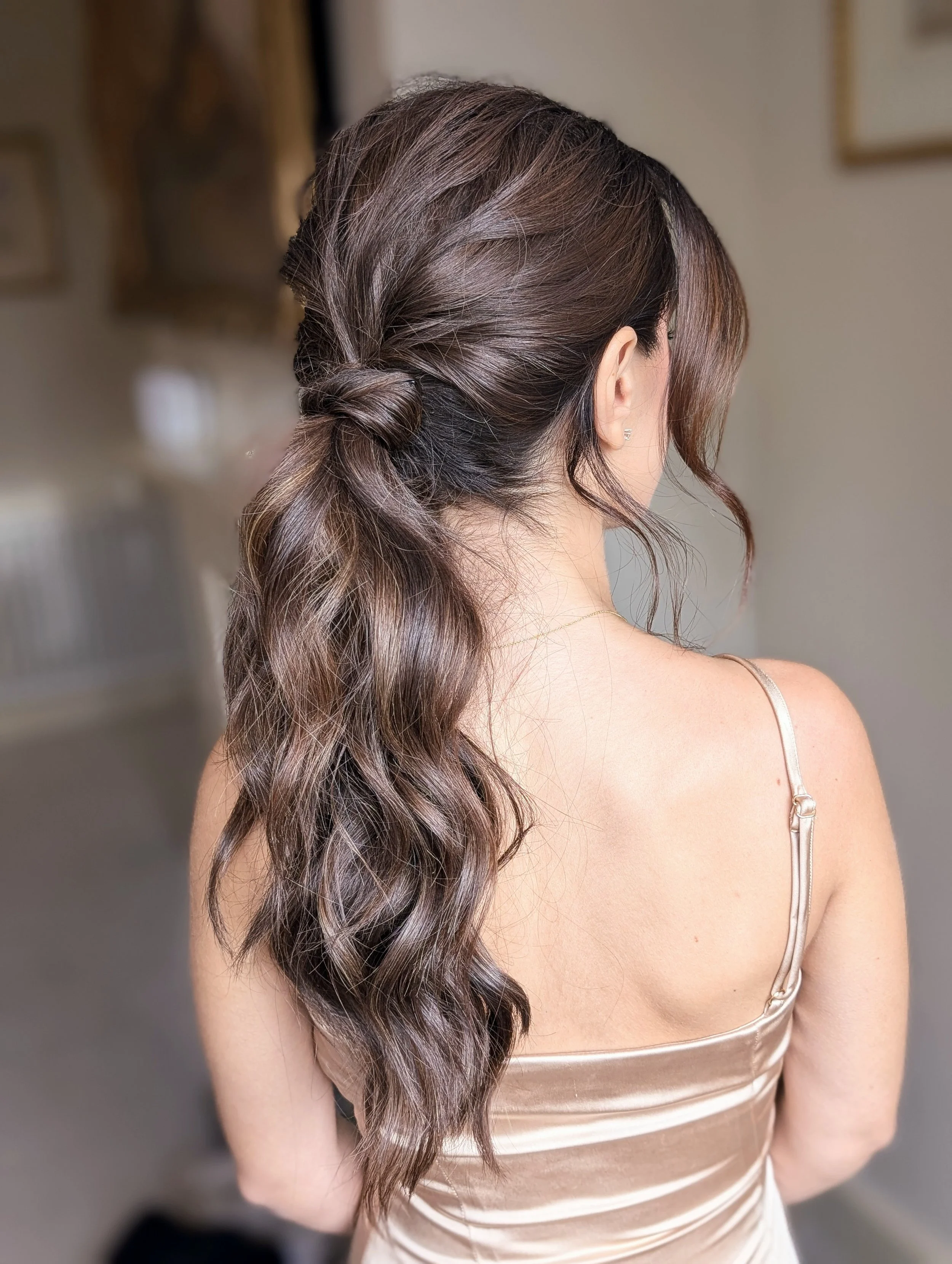 Back view of a woman with wavy brown hair styled in a loose half-updo, wearing a satin spaghetti strap dress.