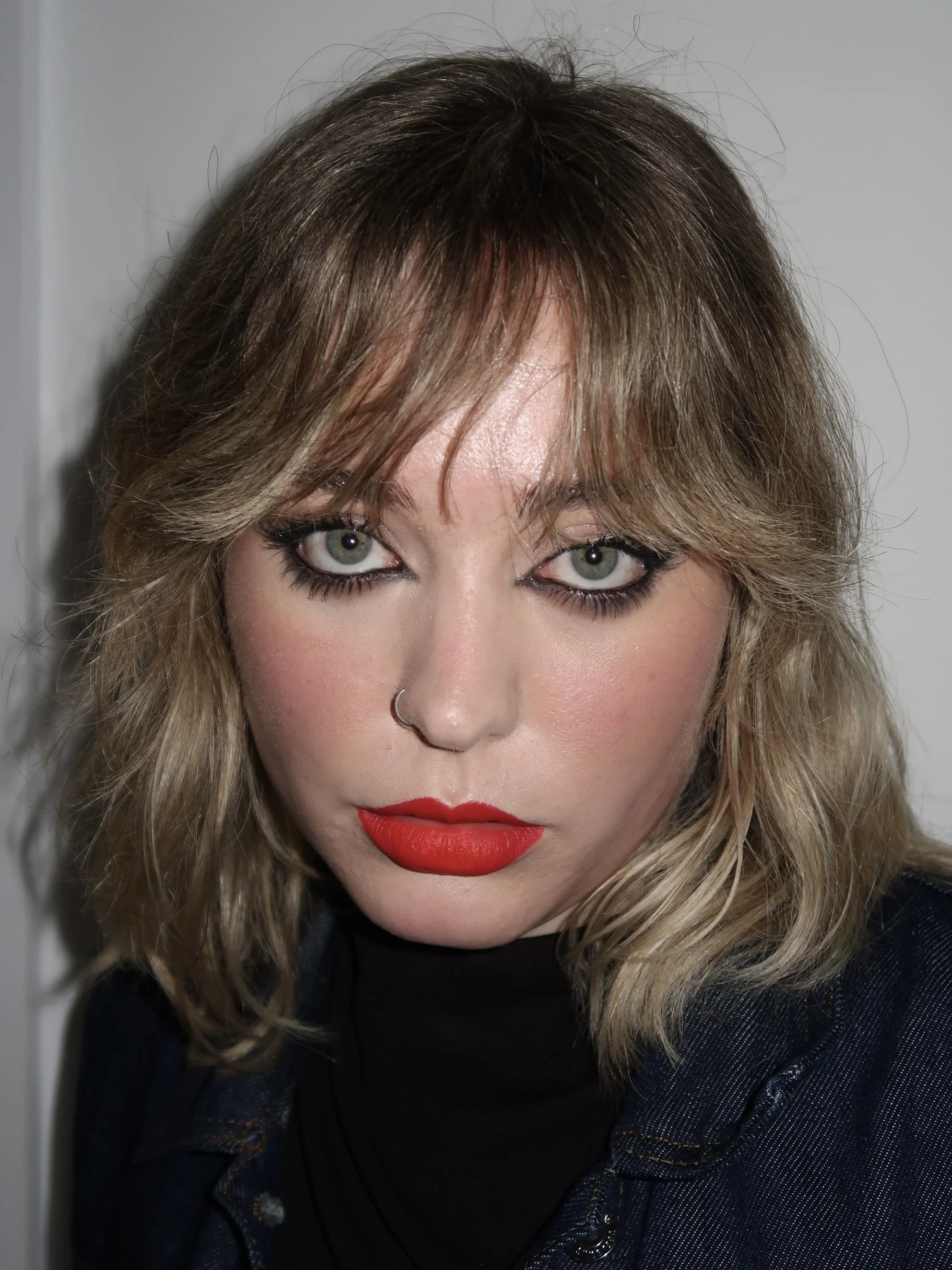 A woman with blonde, wavy hair and light-colored eyes, wearing black eye makeup and red lipstick, with a nose ring, looking directly at the camera.