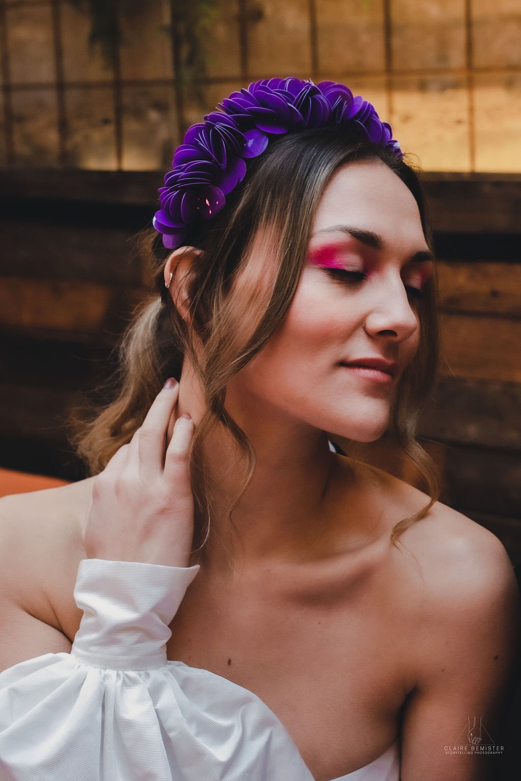 A woman with closed eyes wearing a purple floral headpiece and pink eyeshadow, touching her neck with her left hand, with a wooden wall background.