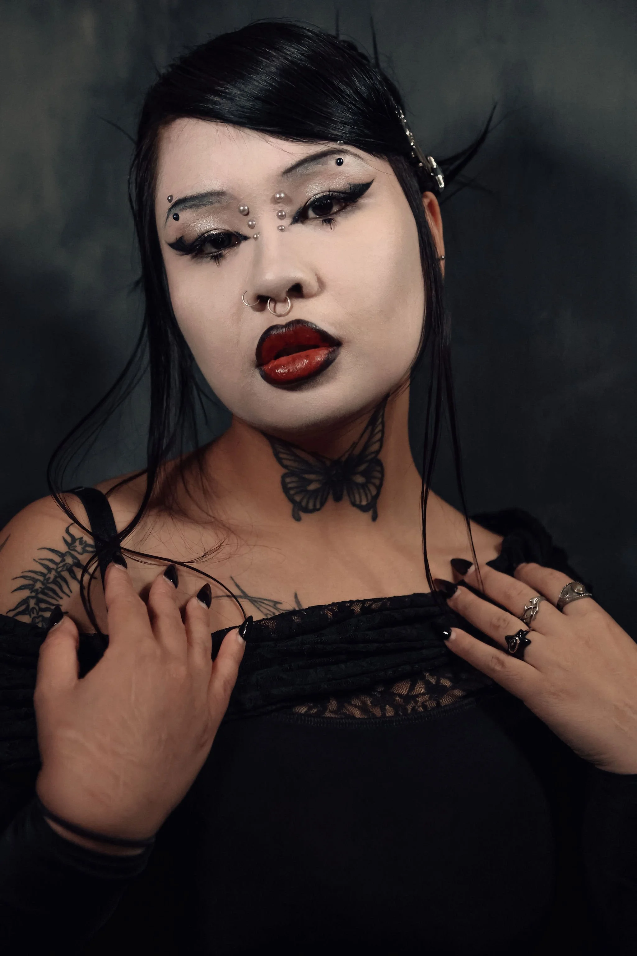 A woman with black hair and gothic makeup, including dark lipstick and dramatic eye makeup, poses against a dark background. She has facial piercings, tattoos on her shoulder and neck, and wears rings and dark clothing with lace details.