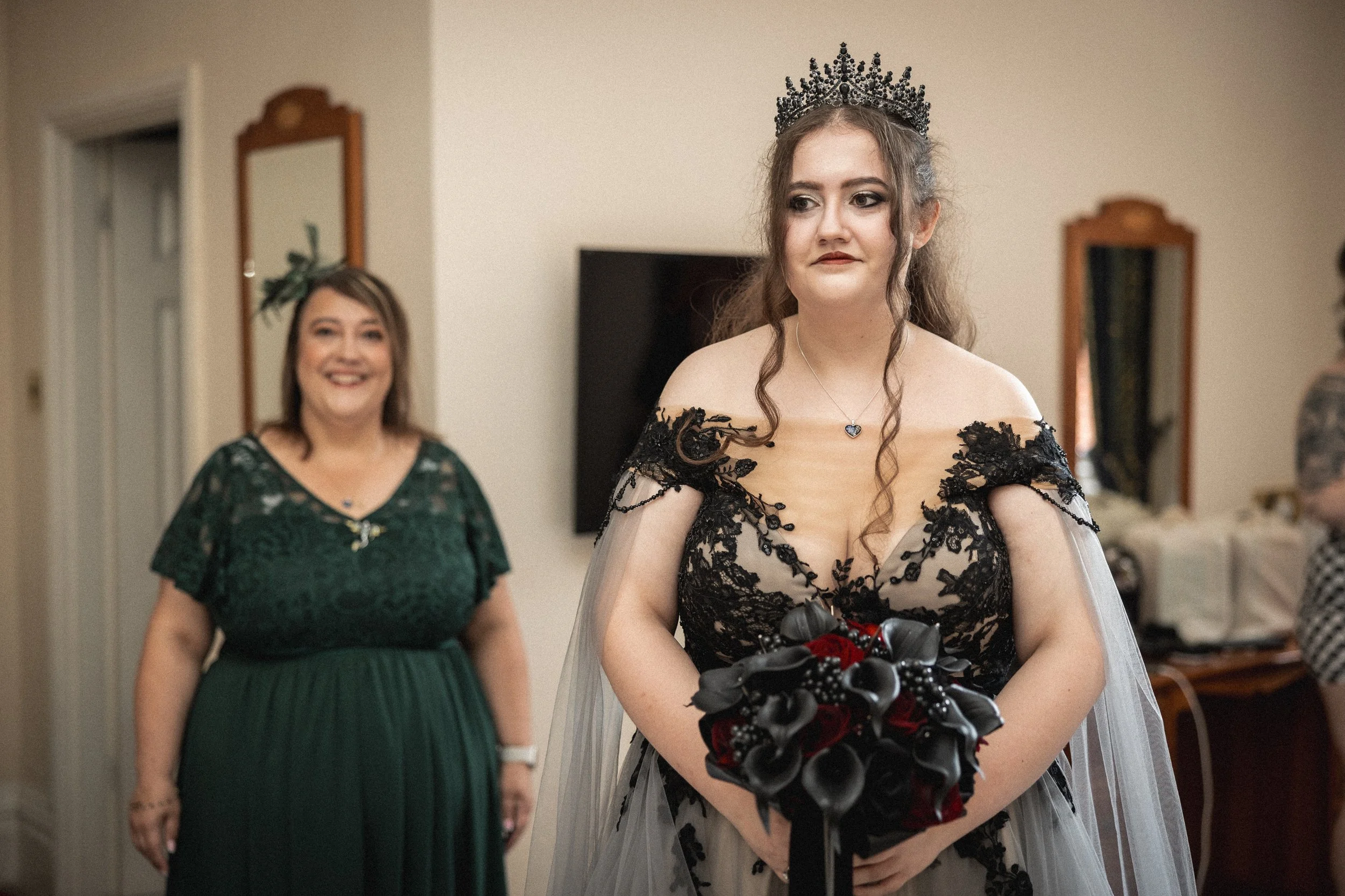 A bride in a black lace wedding gown and black crown holds a bouquet, standing indoors. Behind her, a woman in a green dress smiles.
