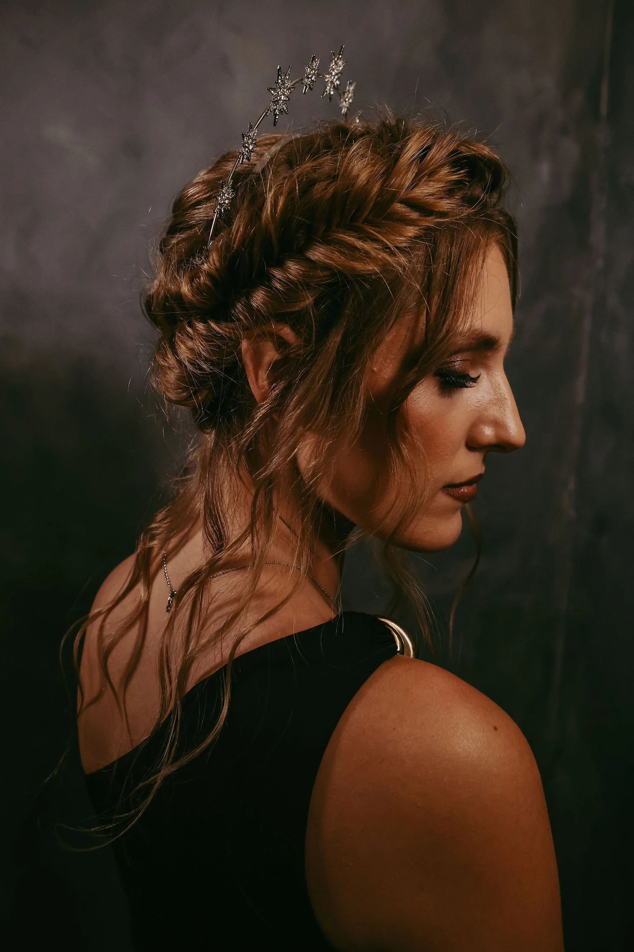 A woman with styled wavy hair wearing a silver star-shaped headband, black sleeveless top, and a delicate necklace, facing sideways against a dark background.