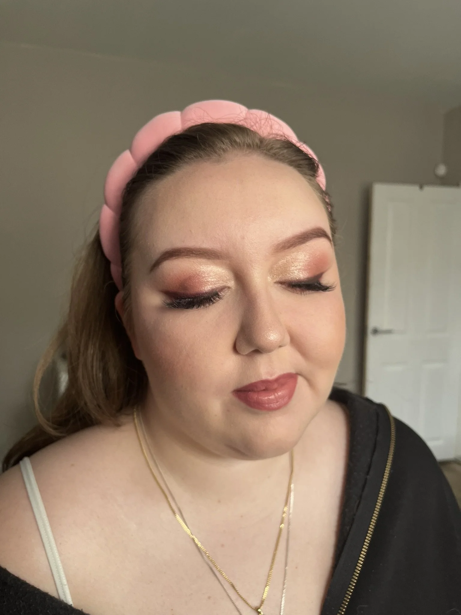 A woman with light skin and brown hair wearing a pink headband, gold necklaces, and makeup with gold and pink eyeshadow, false eyelashes, and nude lipstick, looking down with her eyes closed.