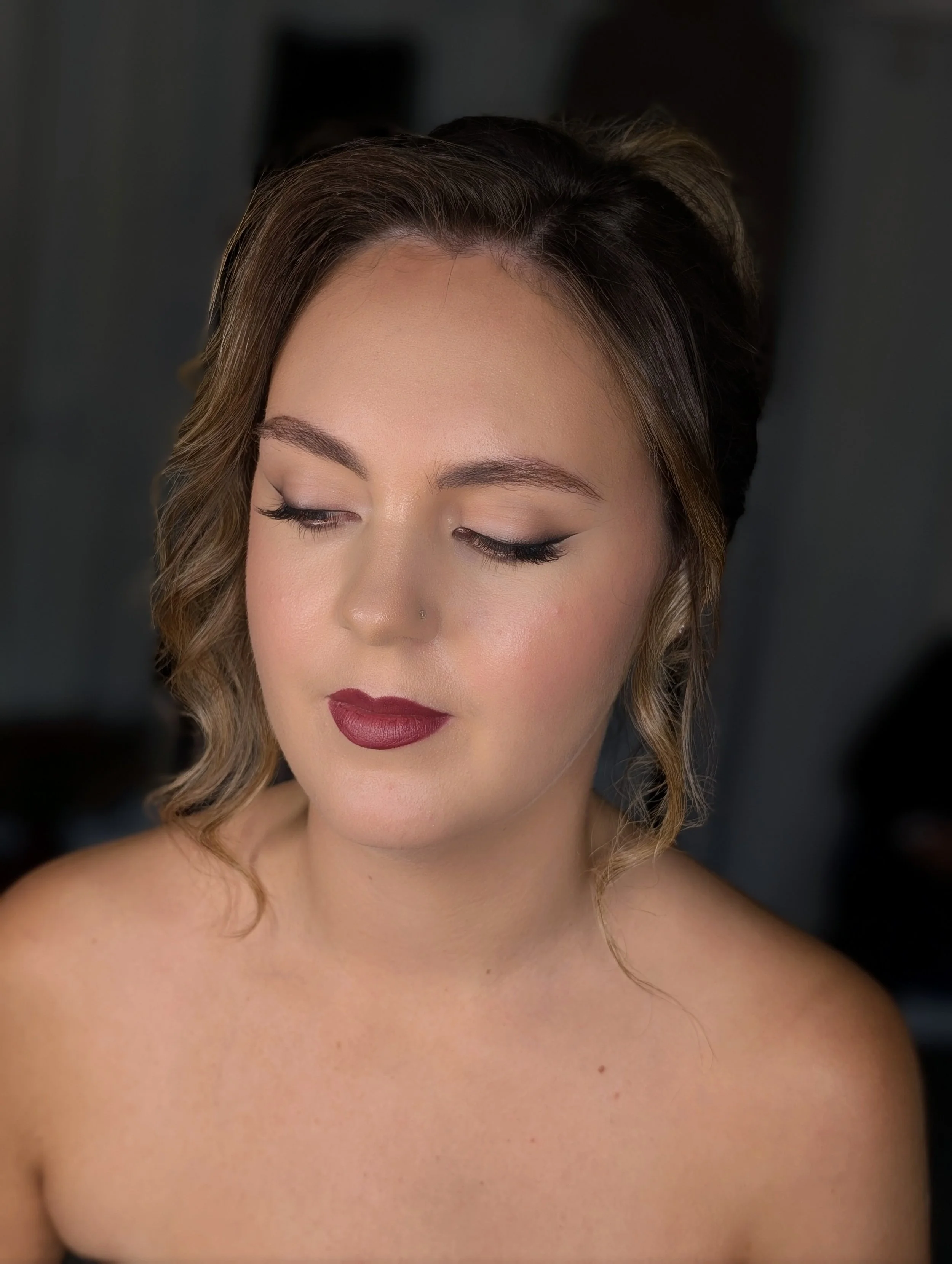Close-up of a young woman with styled hair, makeup, and a nose piercing, looking downward with her eyes closed.