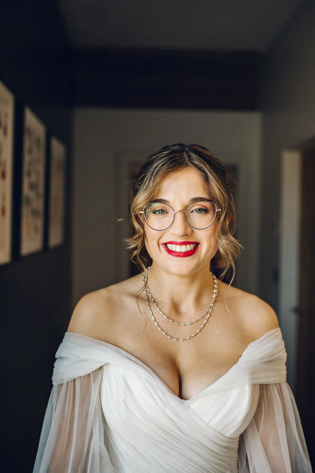 A smiling woman with glasses, wearing red lipstick, a white off-shoulder dress, and layered pearl necklaces, standing indoors.