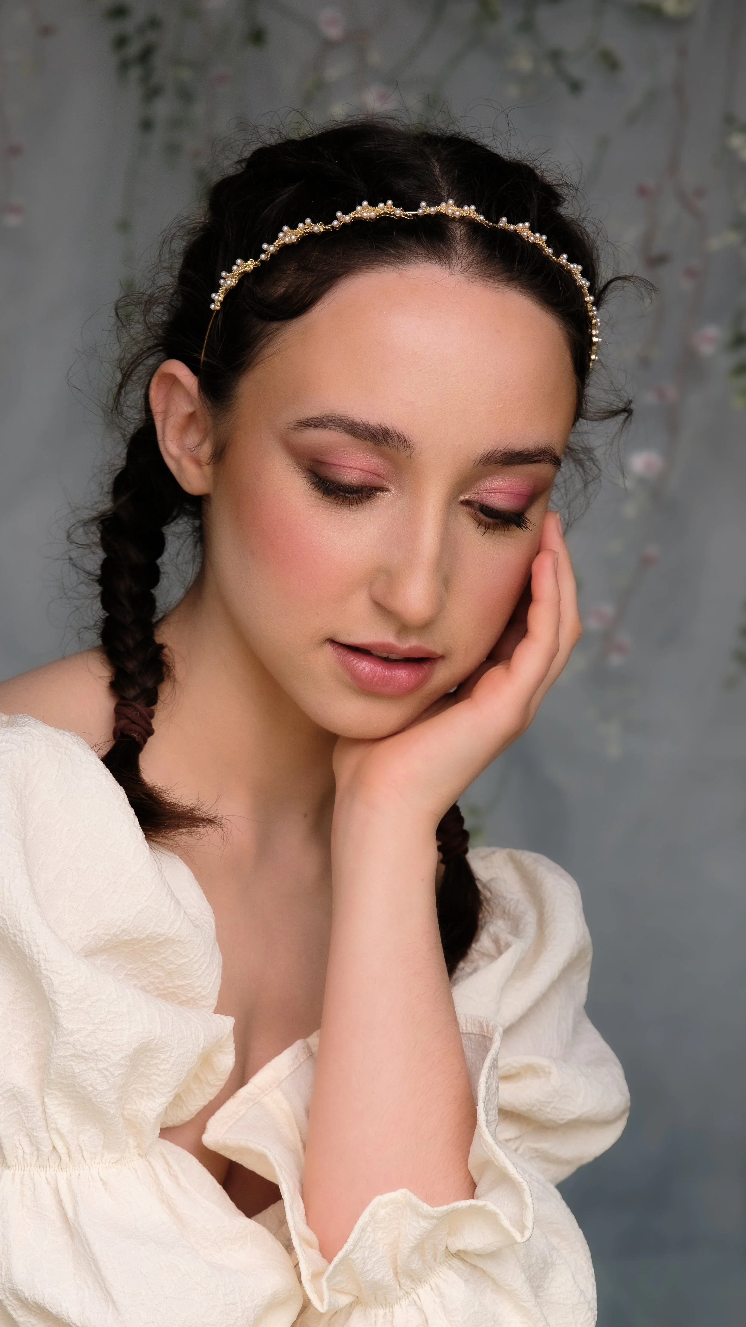 A young woman with braided hair, wearing a pearl headband, has her eyes closed and touches her face with her hand, dressed in an off-white puff-sleeve top, with a blurred floral background.