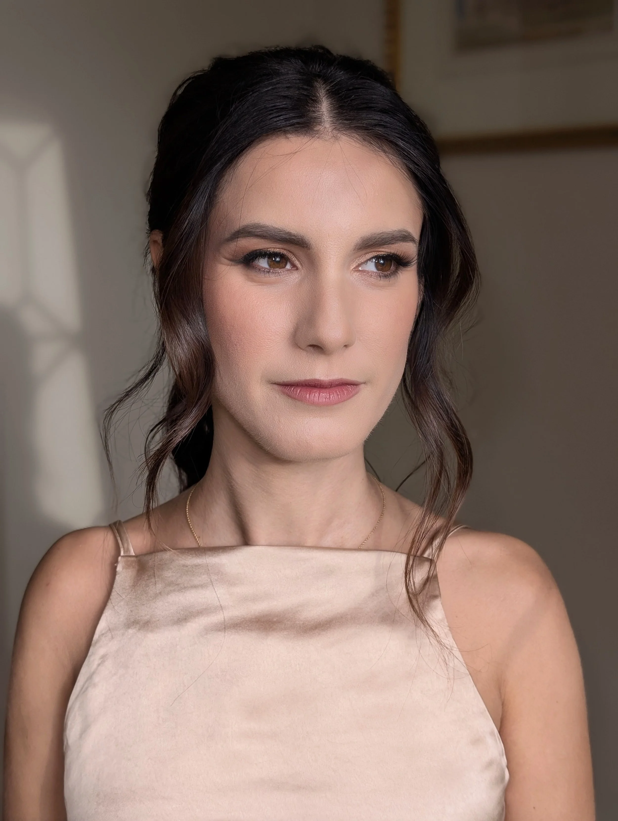 Portrait of a woman with dark hair styled in loose waves, wearing a beige satin sleeveless top, in a softly lit indoor setting.