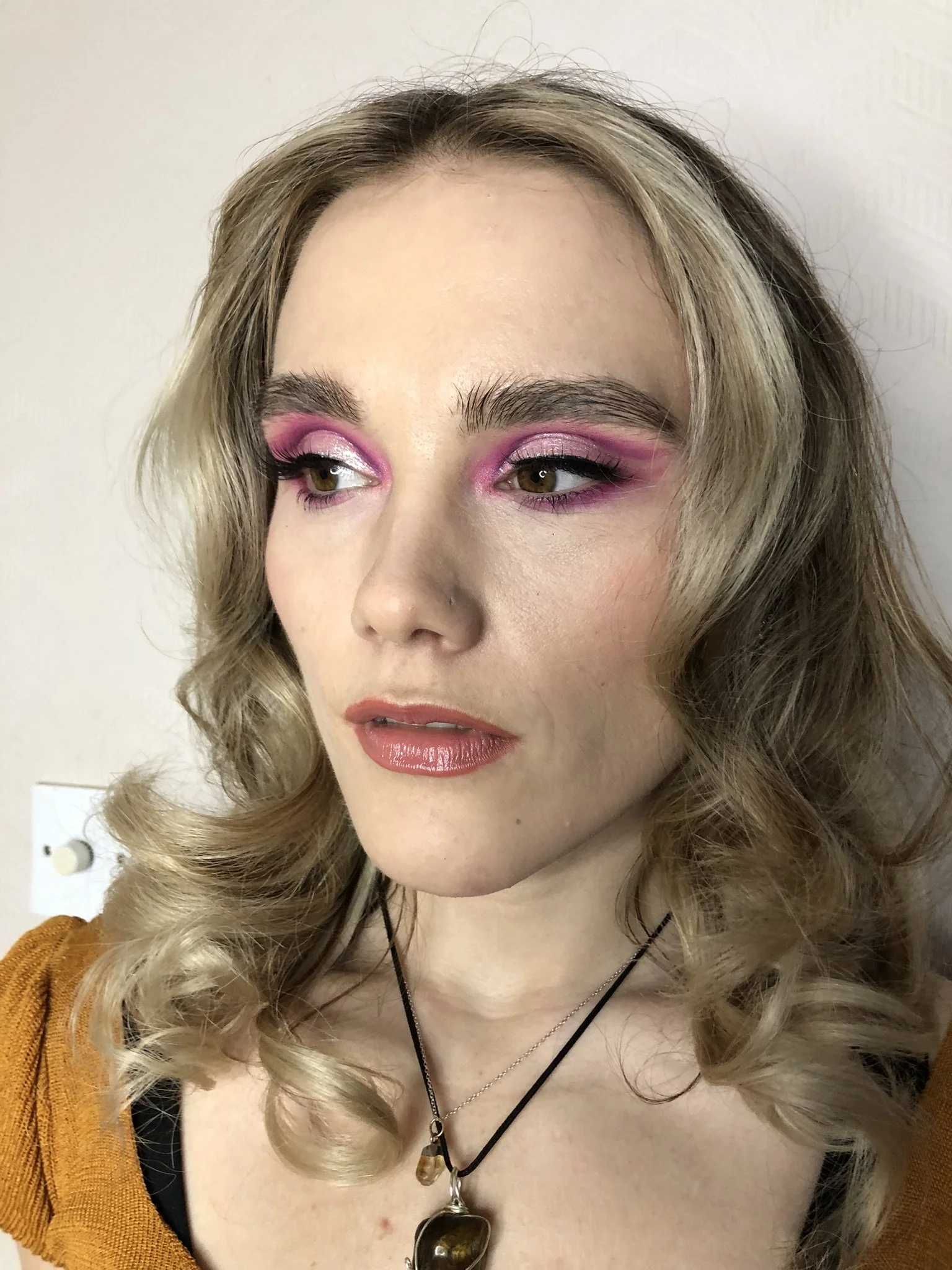 Close-up of a woman with curly blonde hair, pink eyeshadow, and natural-colored lipstick wearing layered necklaces, in front of a plain white wall.