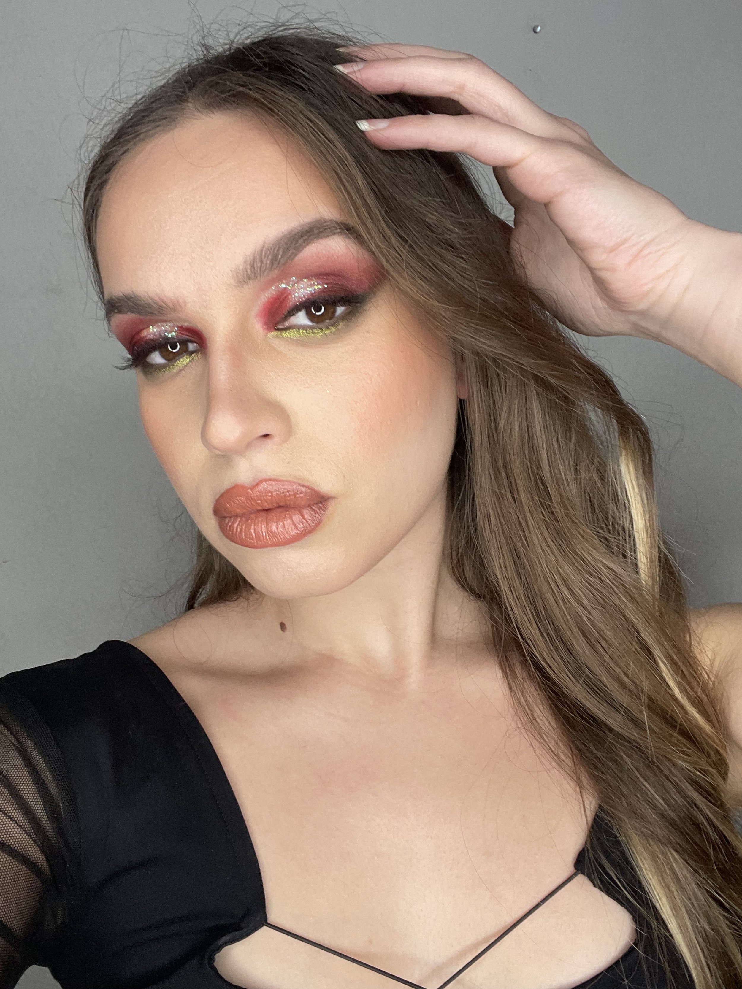 A woman with long brown hair, wearing a black top with sheer sleeves, has makeup including pink and glitter eyeshadow, yellow eyeliner, and peach lipstick. She is holding her hair with her right hand and looking at the camera.