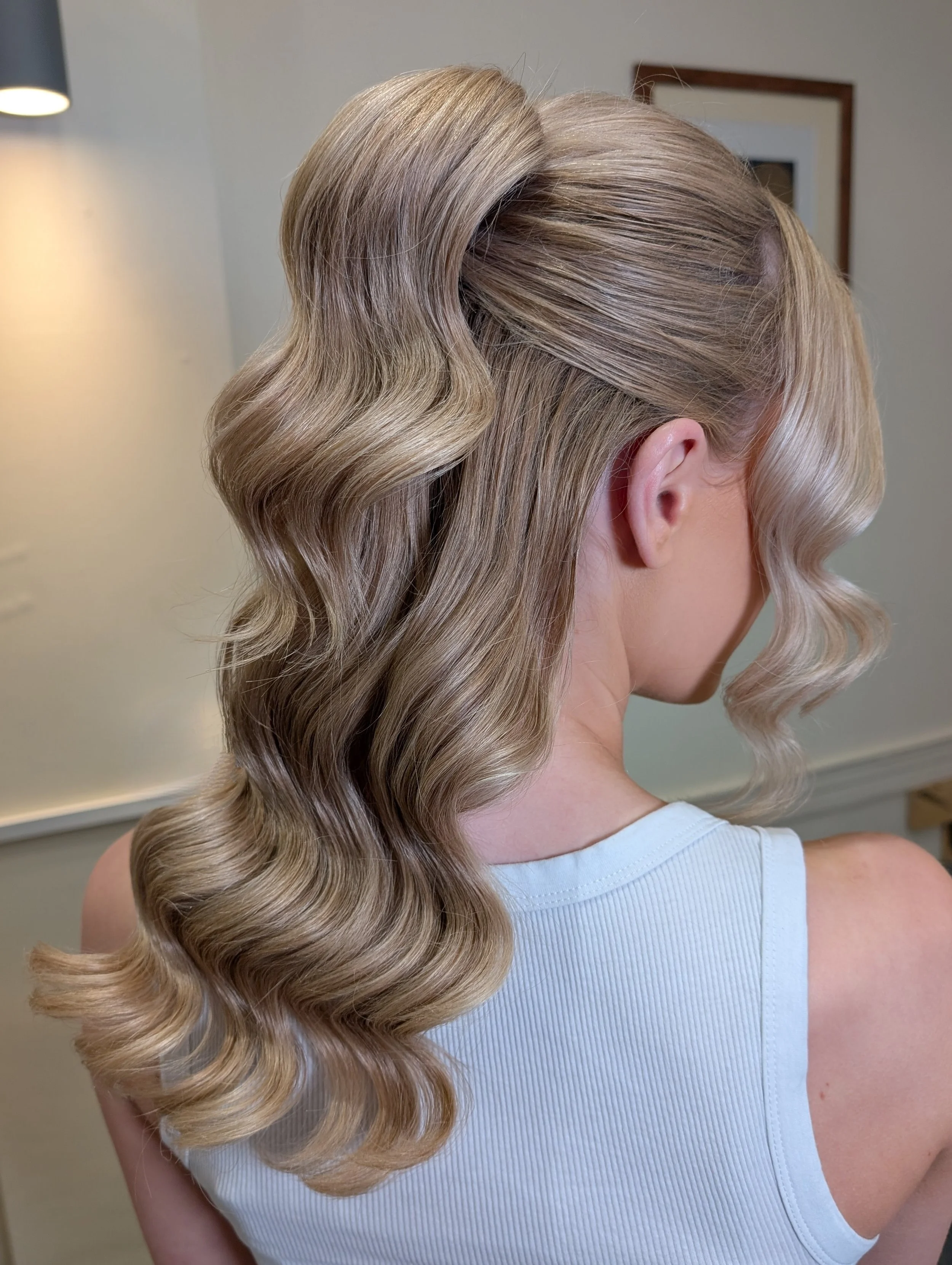 Side view of a woman with styled, wavy blonde hair, wearing a white tank top, indoors.