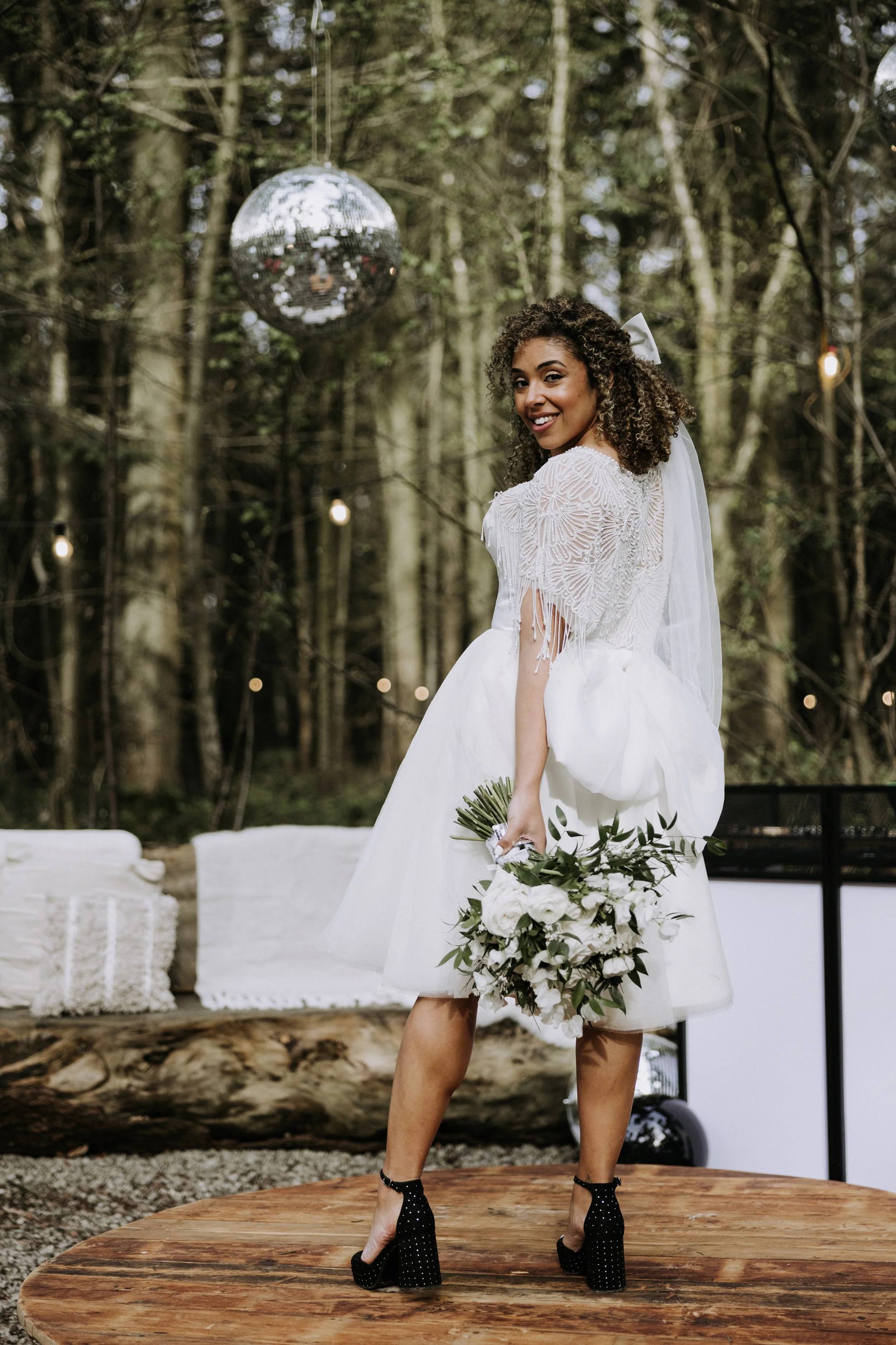 A woman in a wedding dress holding a bouquet, standing on a wooden platform outdoors in a wooded area, with a disco ball hanging above.