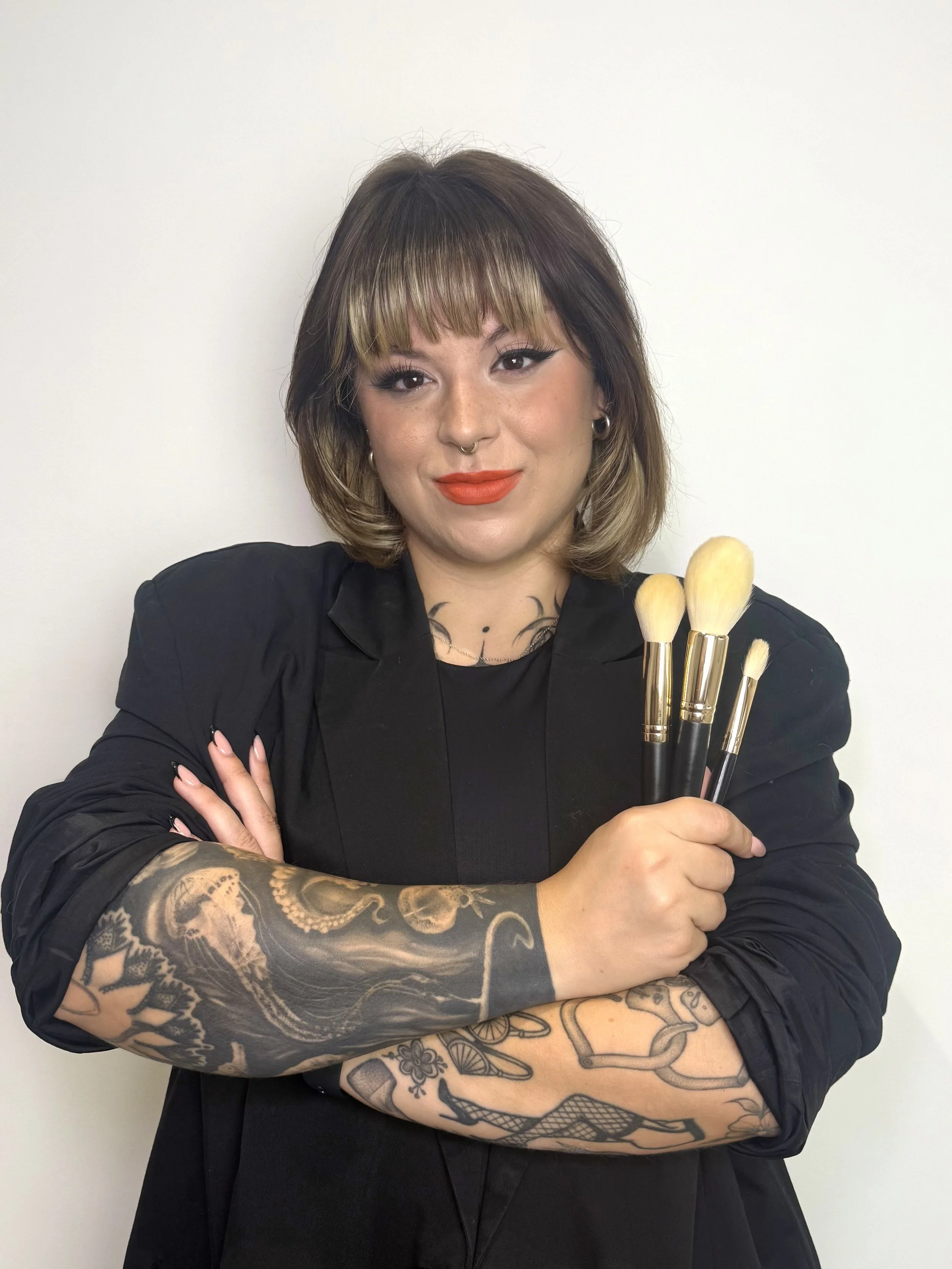 A woman with shoulder-length brown hair, styled with blonde bangs, is holding a set of makeup brushes and smiling. She has tattoos on her arms, winged eyeliner, and is wearing a black top and blazer, standing against a plain white background.