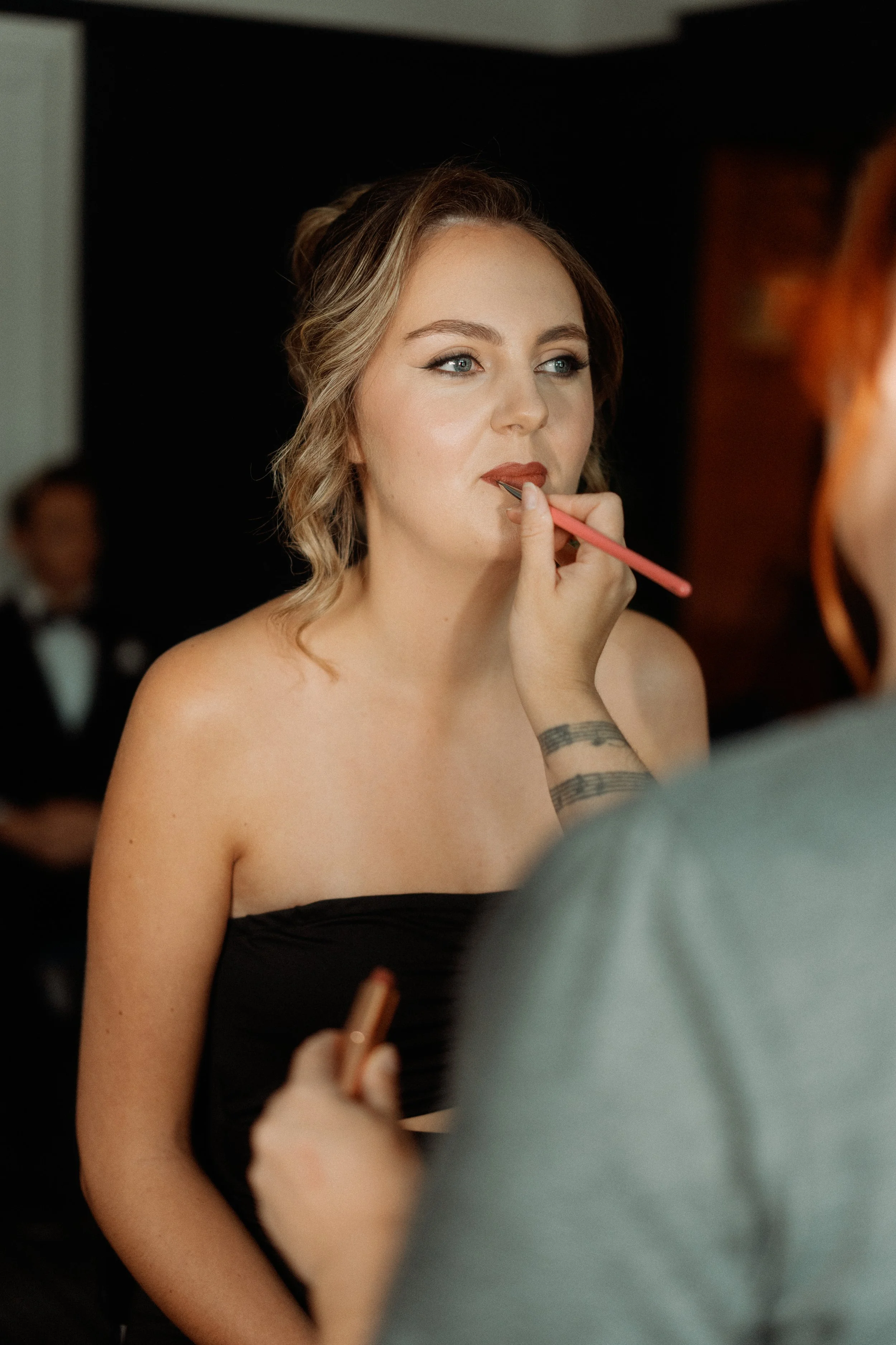 A woman with fair skin and wavy blonde hair in a loose updo is getting her makeup done, with a makeup artist applying lipstick. The woman is wearing a black strapless dress and has blue eyes and makeup, including eyeliner and lipstick.