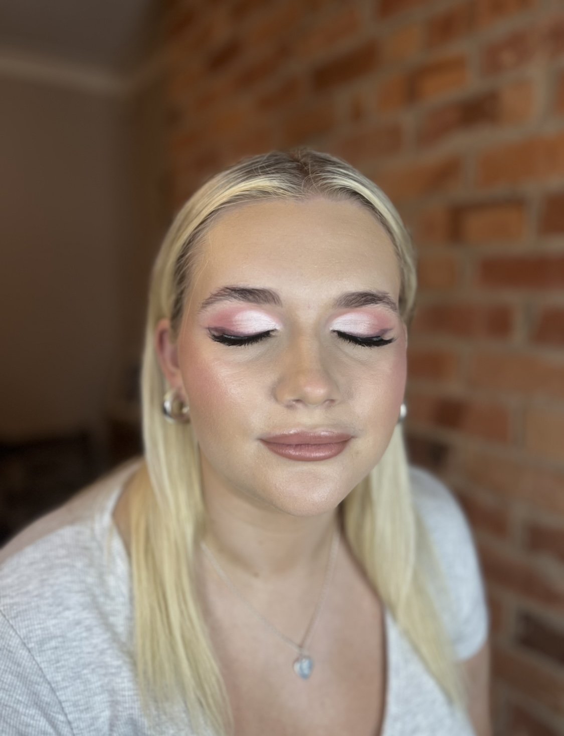 Close-up of a woman with blonde hair, closed eyes showing pink and white eye makeup, wearing silver earrings and a pendant necklace, in front of a brick wall.