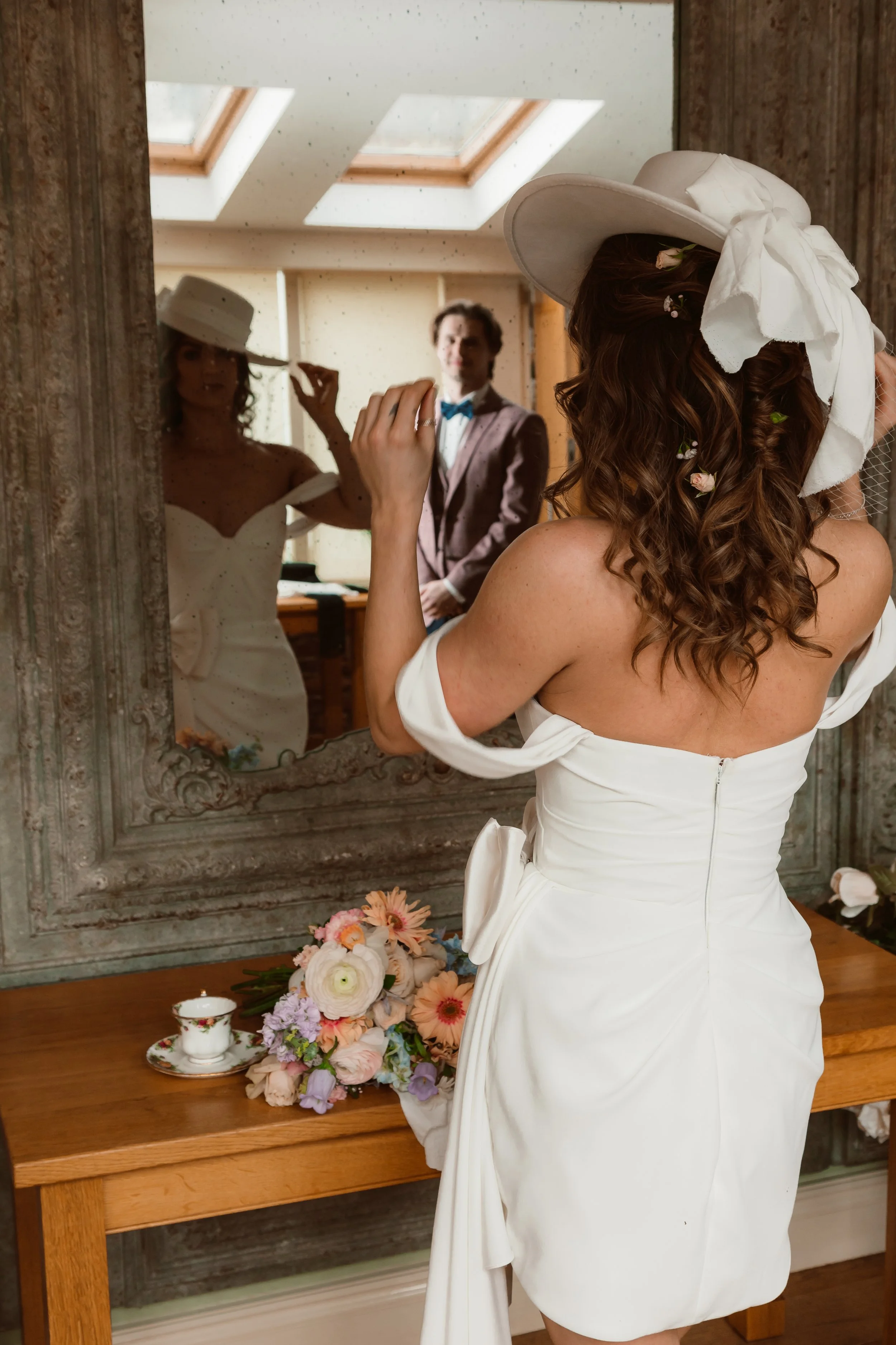 A woman in a white wedding dress and wide-brimmed hat is looking at herself in a mirror, with a bouquet of flowers on a table in front of her. A man in a suit and bow tie stands in the background, reflected in the mirror.