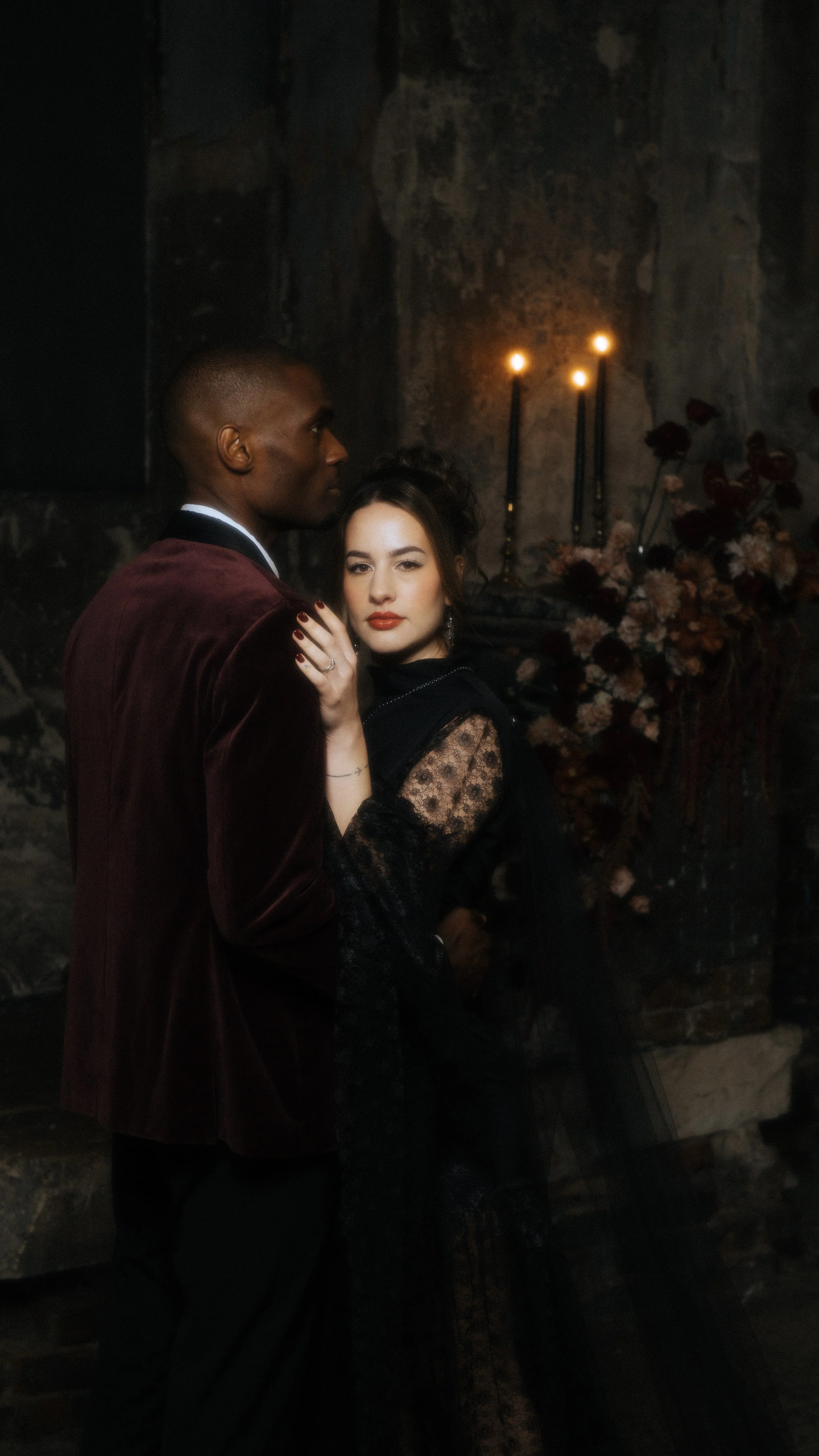A darkly lit scene with a man and woman in elegant attire. The man wears a maroon velvet blazer, and the woman is dressed in a black lace dress. They stand close together, with the woman's hand on the man's shoulder, and a romantic ambiance with candles and flowers in the background.