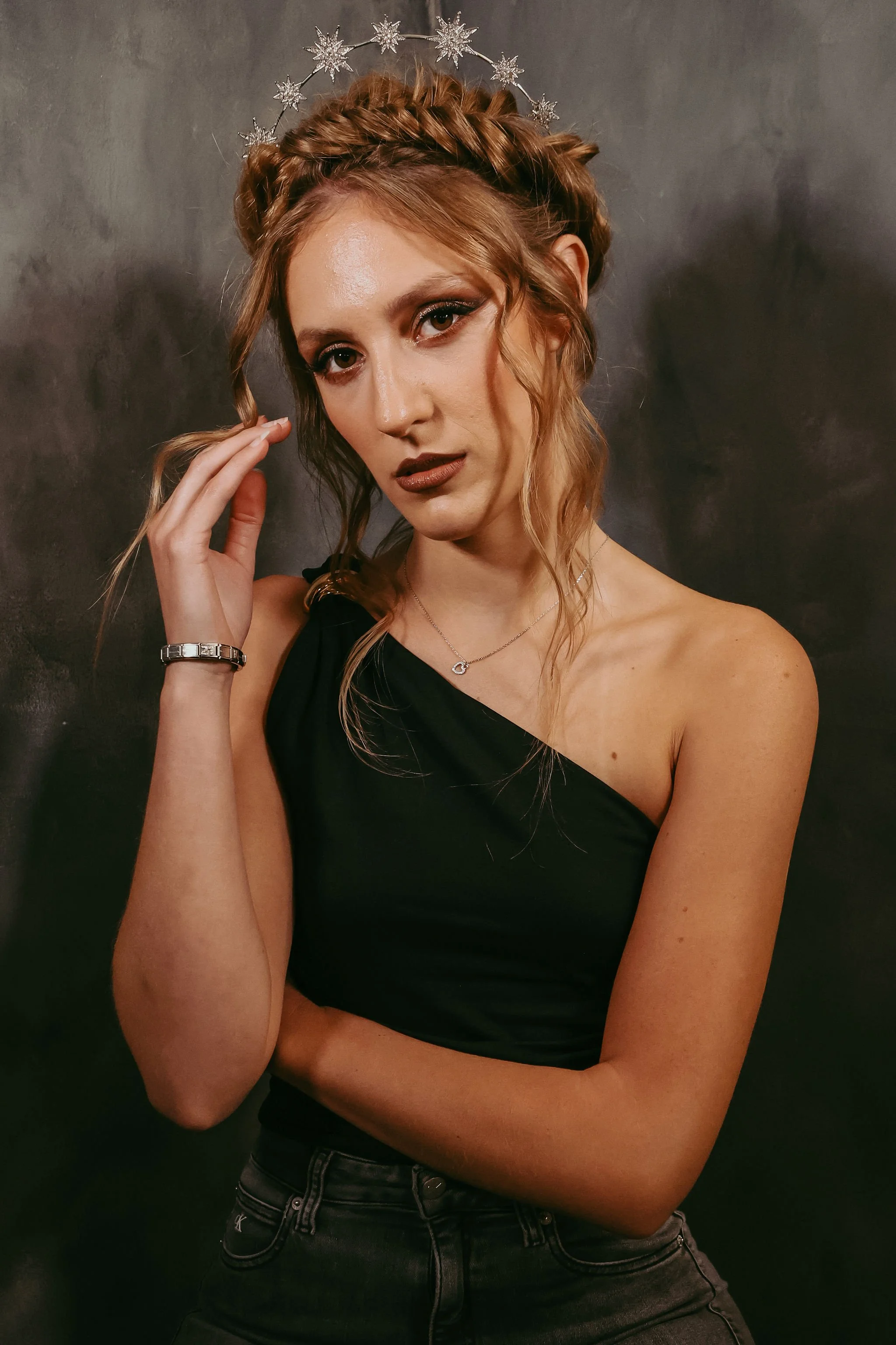 A woman with wavy blonde hair wearing a black one-shoulder top and black jeans, with a silver bracelet, necklace, and a silver snowflake halo crown, posing against a dark background.