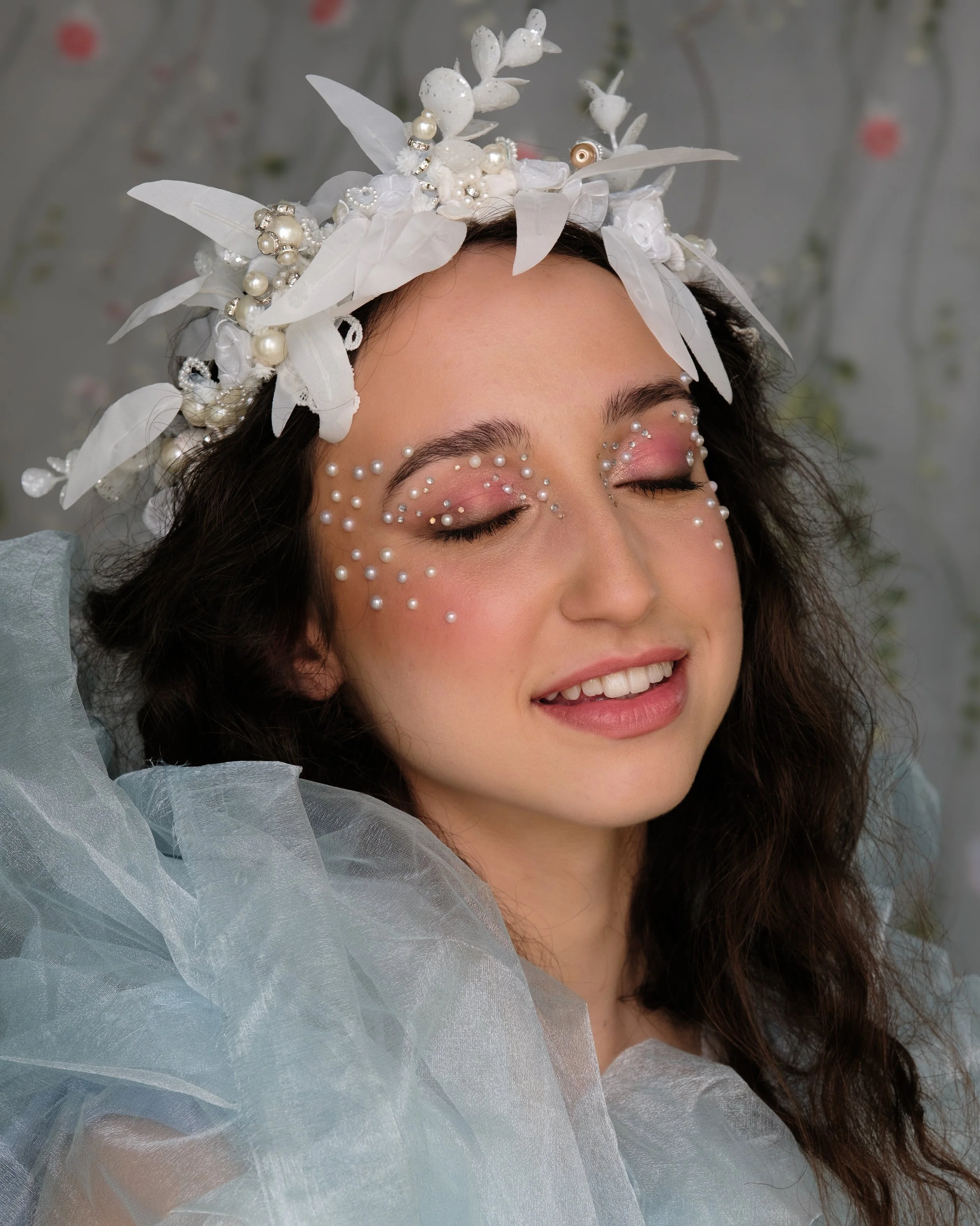 A woman with long, curly brown hair and closed eyes, wearing a white floral headpiece decorated with pearls and beads, with makeup featuring pearl embellishments around her eyes, pink eyeshadow, and soft pink lipstick, dressed in a sheer, light blue 