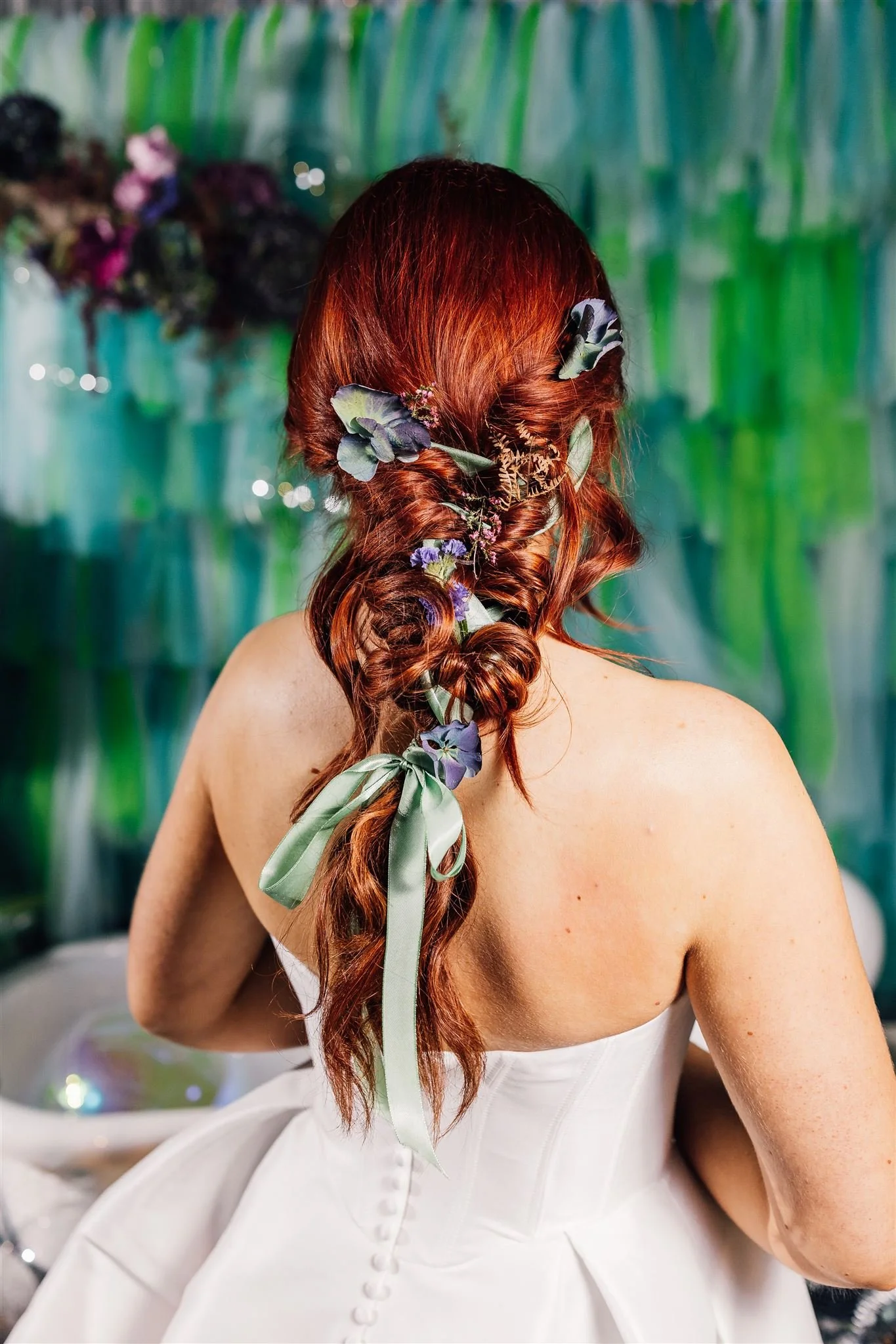 Back view of a woman with long red hair styled in a loose braid, adorned with purple flowers and green ribbon, wearing a white off-the-shoulder dress at a decorated event.