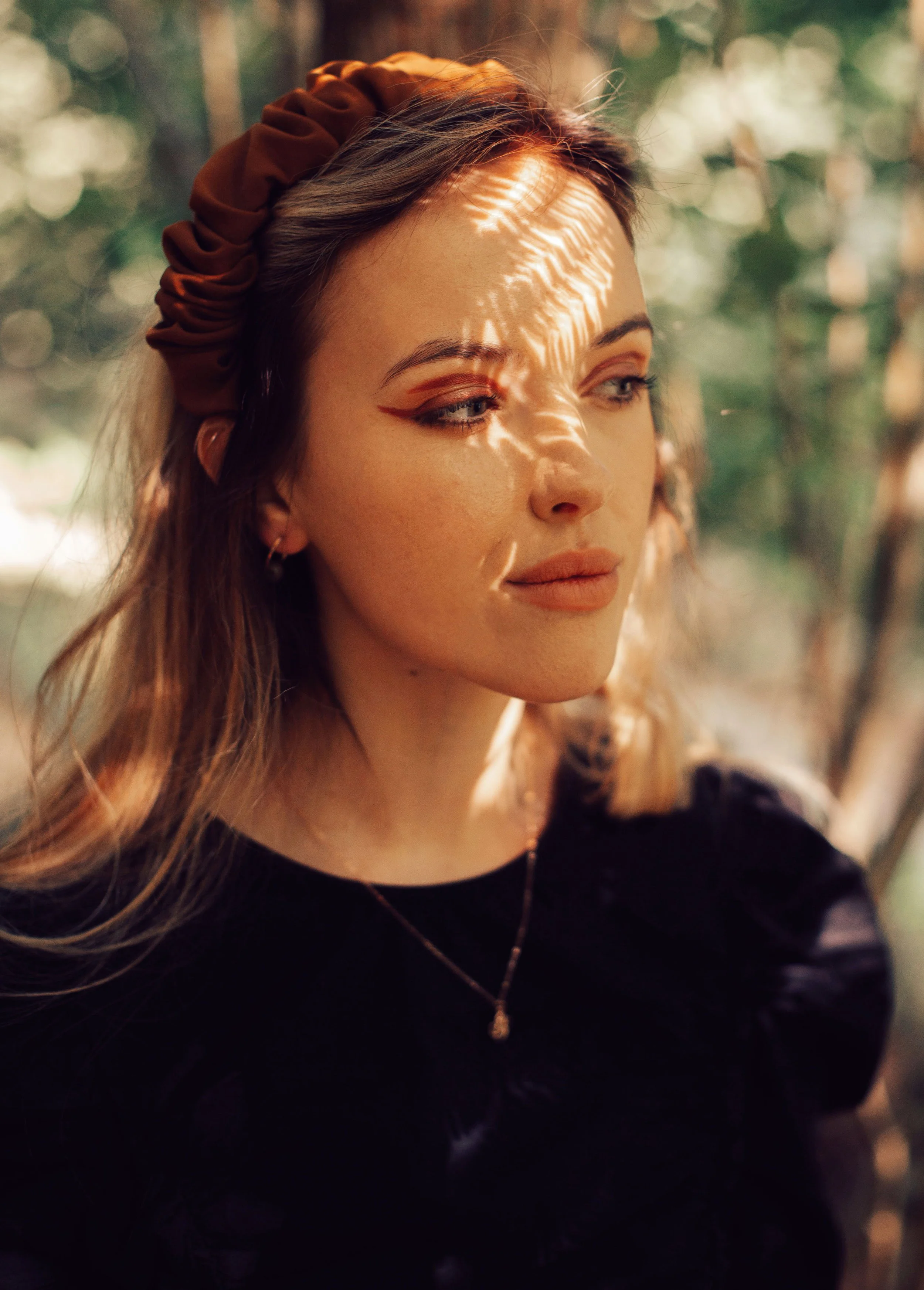 Close-up of a woman with light skin and long, wavy hair, wearing a brown headband, black top, and a gold necklace, with sunlight filtering through leaves creating shadow patterns on her face, outdoors.