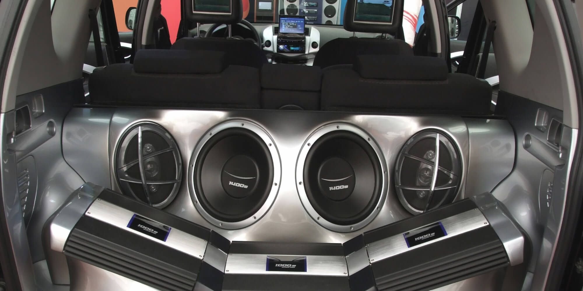 The image shows a large car audio system installed in the trunk of a vehicle, featuring three big subwoofers and two amplifiers, all mounted on a metallic panel.