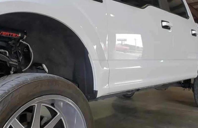 Part of a white vehicle with the front wheel removed, showing suspension components, in an indoor setting.