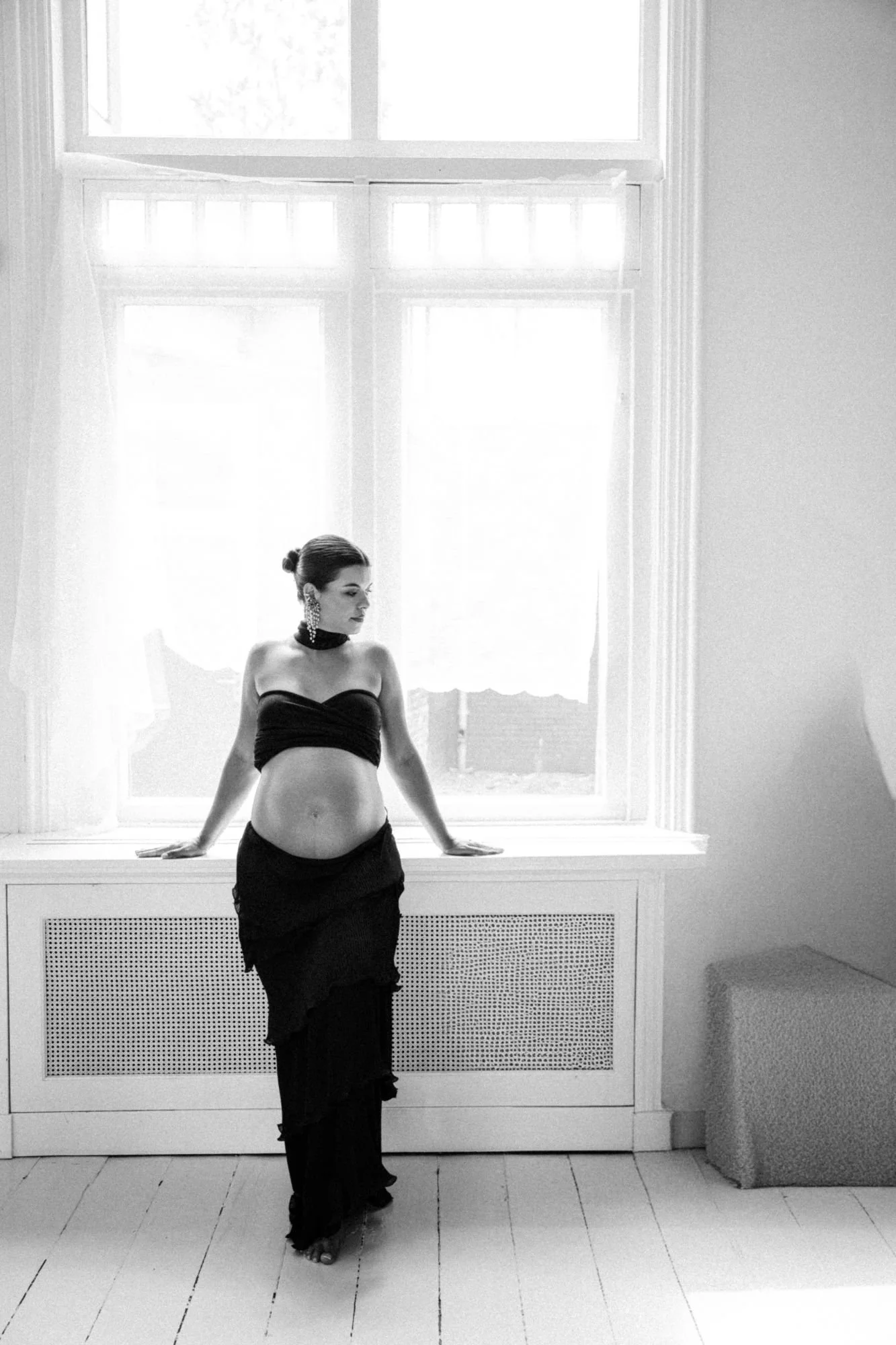 Black-and-white portrait of a pregnant woman in a strapless black dress standing by a bright window.