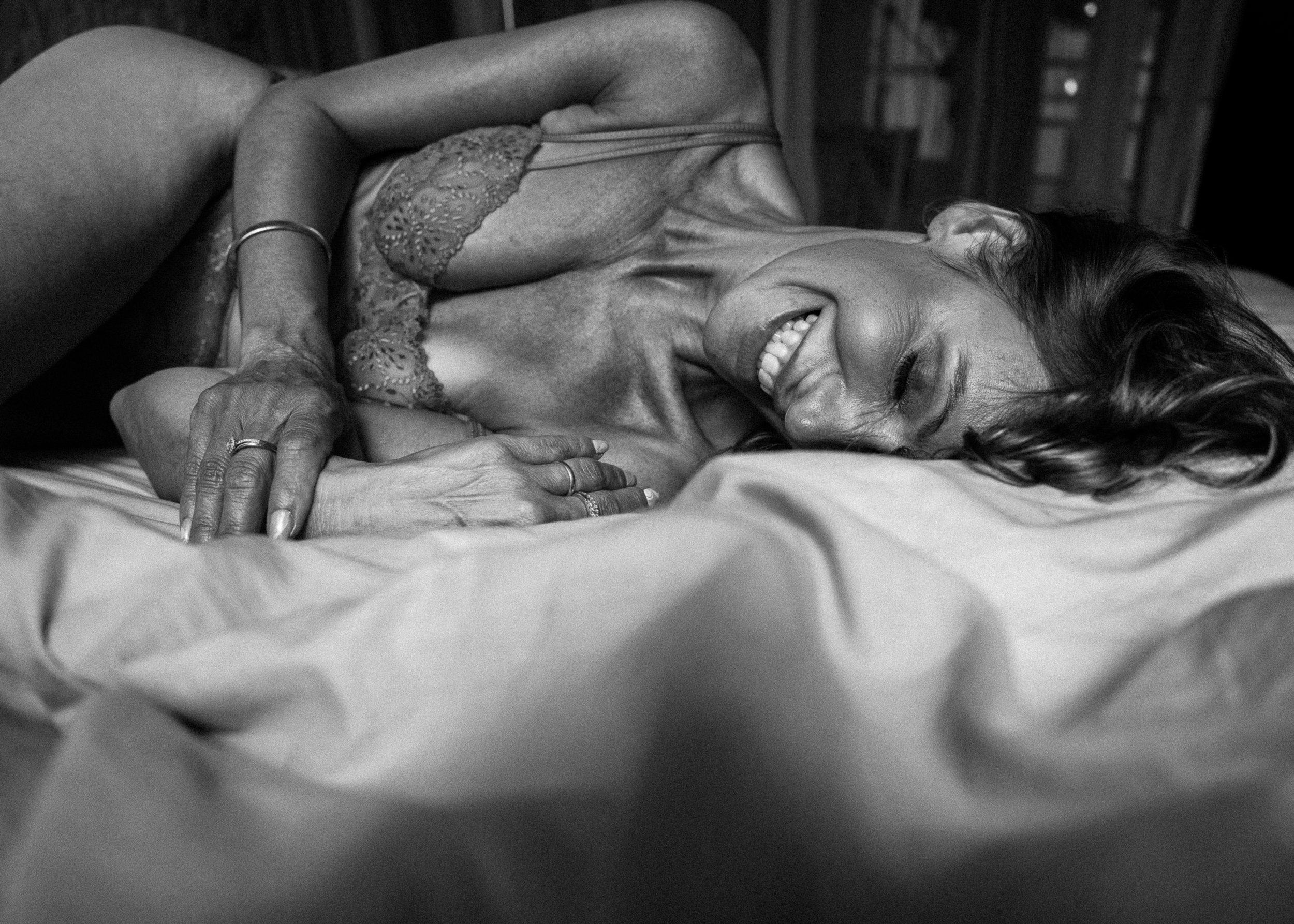 Een glimlachende vrouw die op een bed ligt met gesloten ogen, gekleed in een kanten nachtjurk, in een zwart-witfoto.