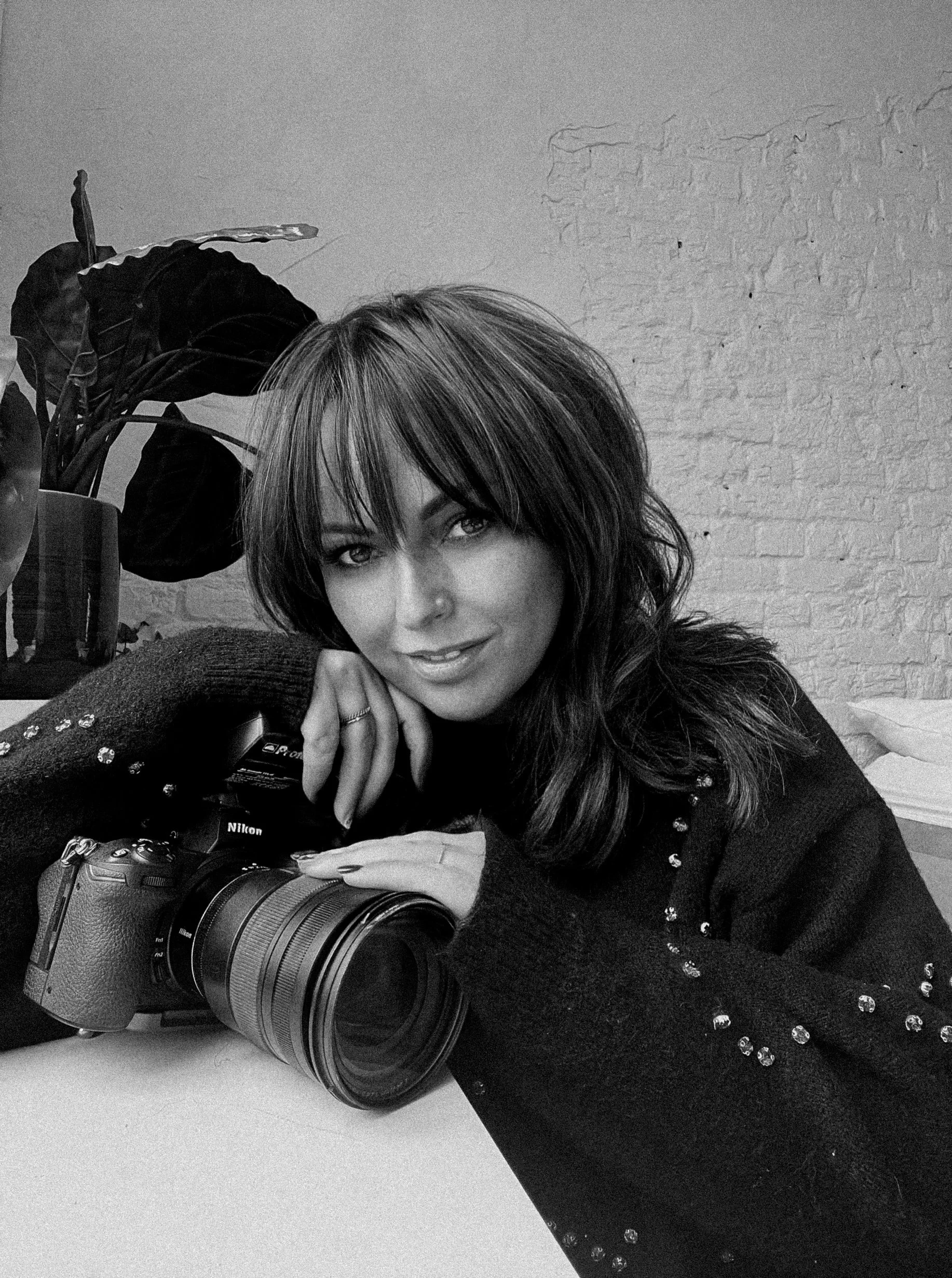 Portret van Wendy De Craemer, fotografe bij ZOOM Fotoatelier, zittend met haar Nikon-camera, zachte blik en glimlach, in een lichte studio-setting.