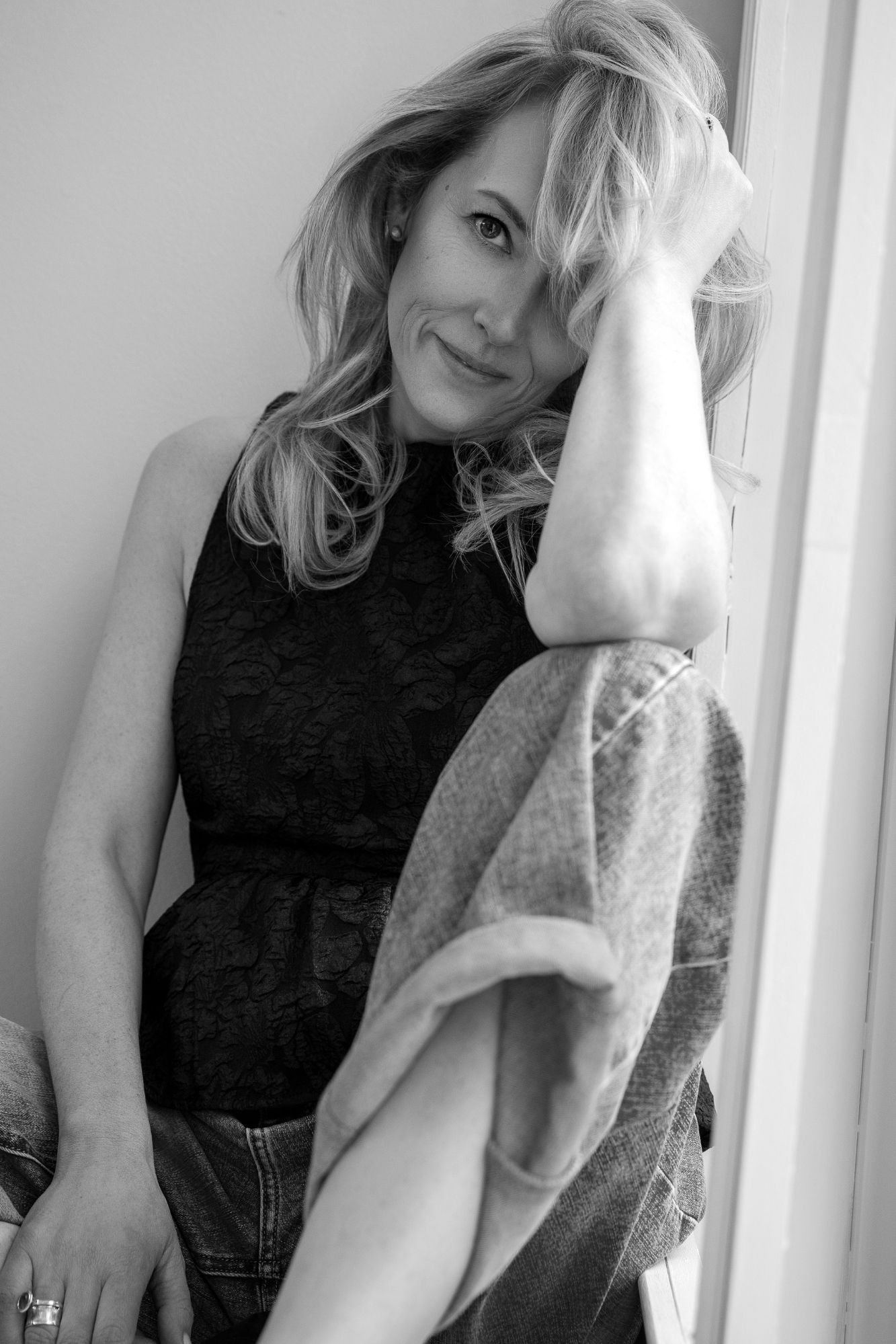 Black-and-white portrait of a smiling woman with wavy hair sitting by a window in a black lace top and jeans.