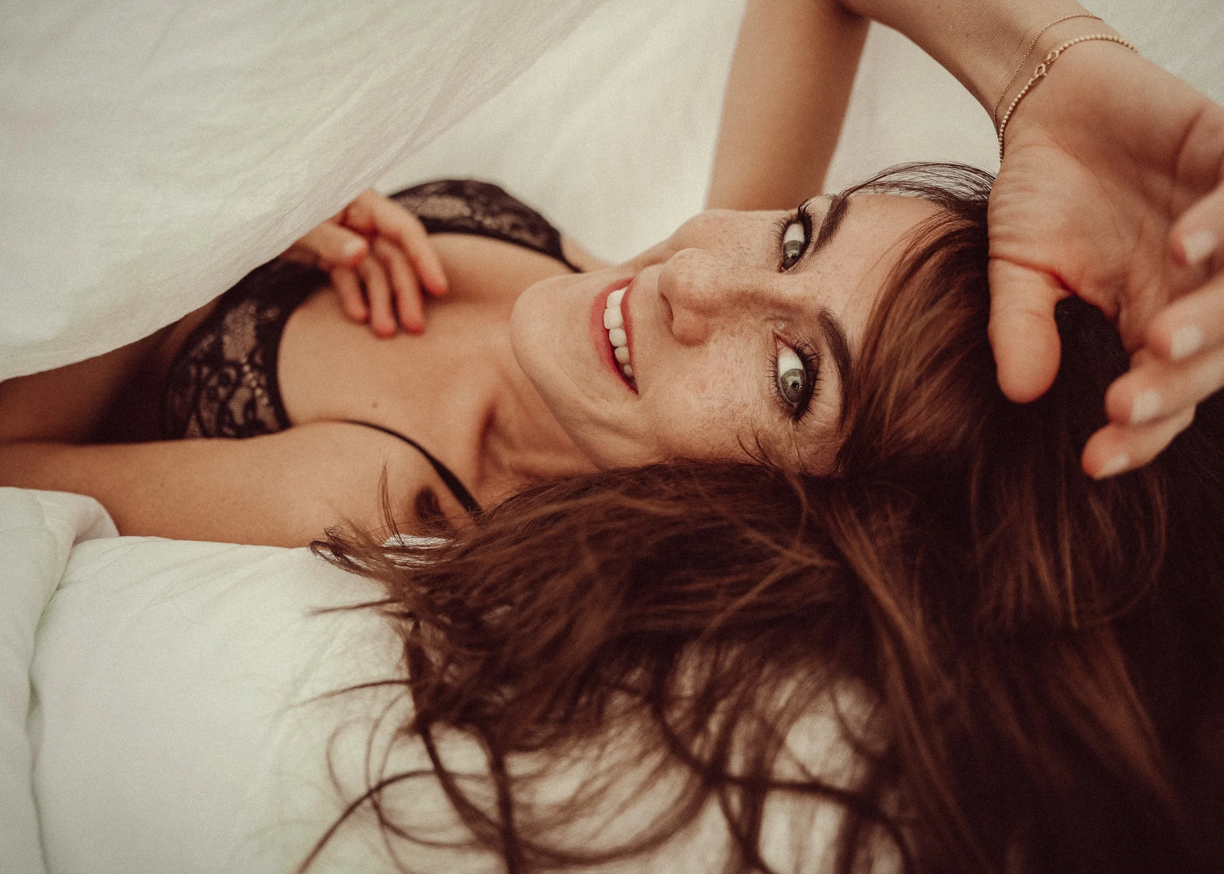 Een vrouw met lang rood haar die op een bed ligt, glimlachend, gekleed in zwarte kanten lingerie. BOUDOIR