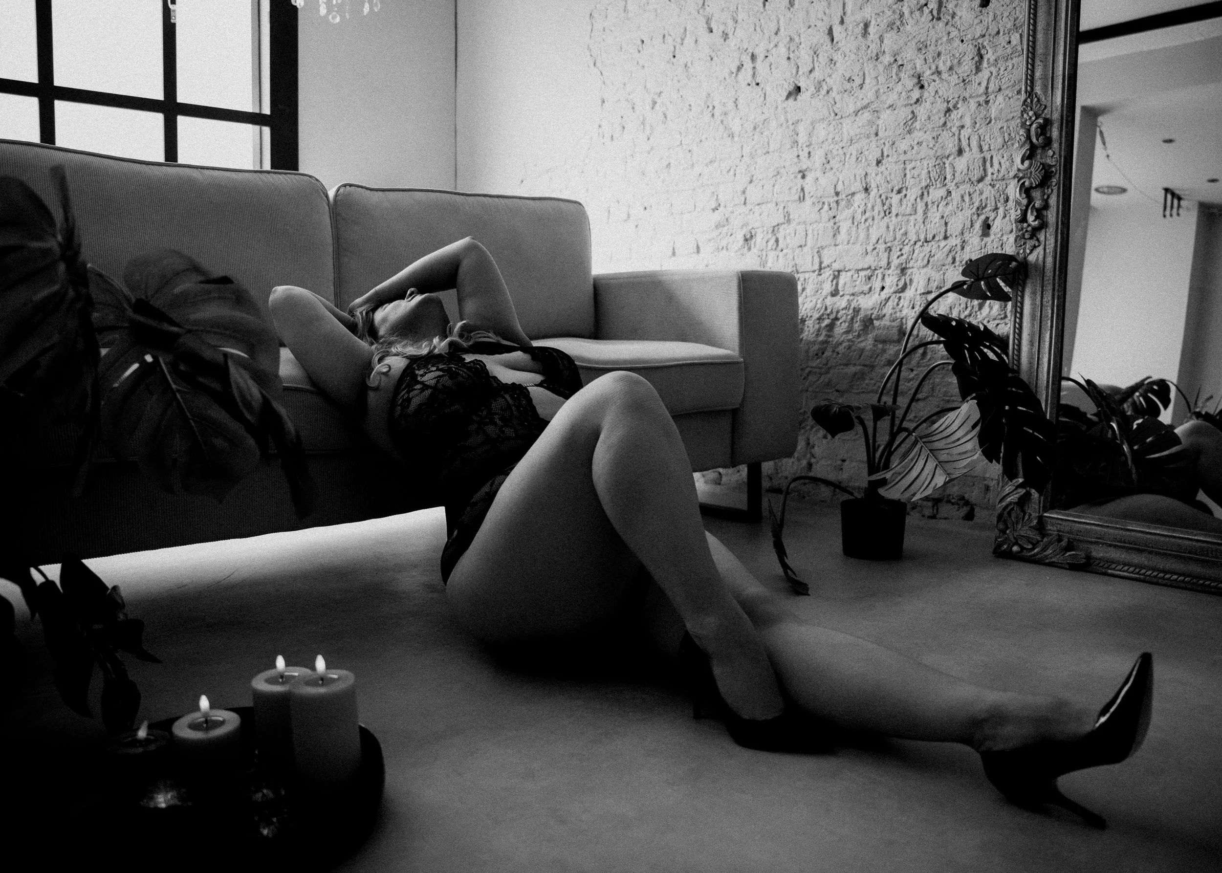Zwart-witfoto van een vrouw die op de vloer ligt en tegen een sofa leunt, gekleed in kanten lingerie en hoge hakken, met kaarsen en planten in een modern ingerichte, industrieel ogende ruimte.