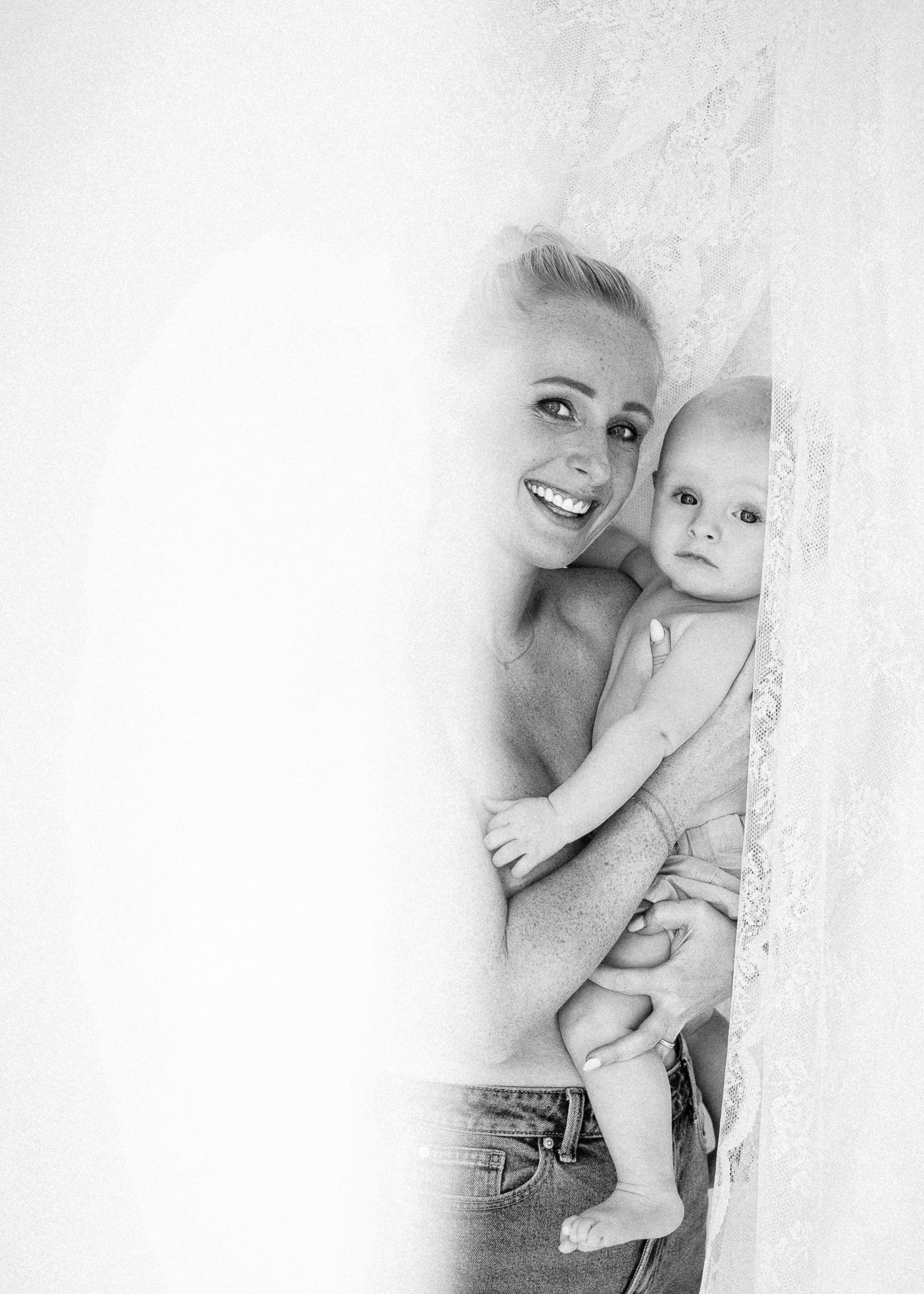A black and white photo of a woman holding a baby, both peeking out from behind a lace curtain. The woman is smiling, and the baby looks curious.