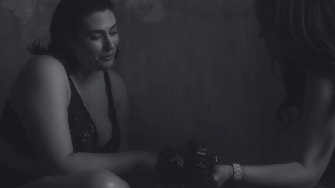 Twee vrouwen die tegenover elkaar zitten en glimlachen; één houdt een camera vast — een warm moment in een donkere, rustige setting.