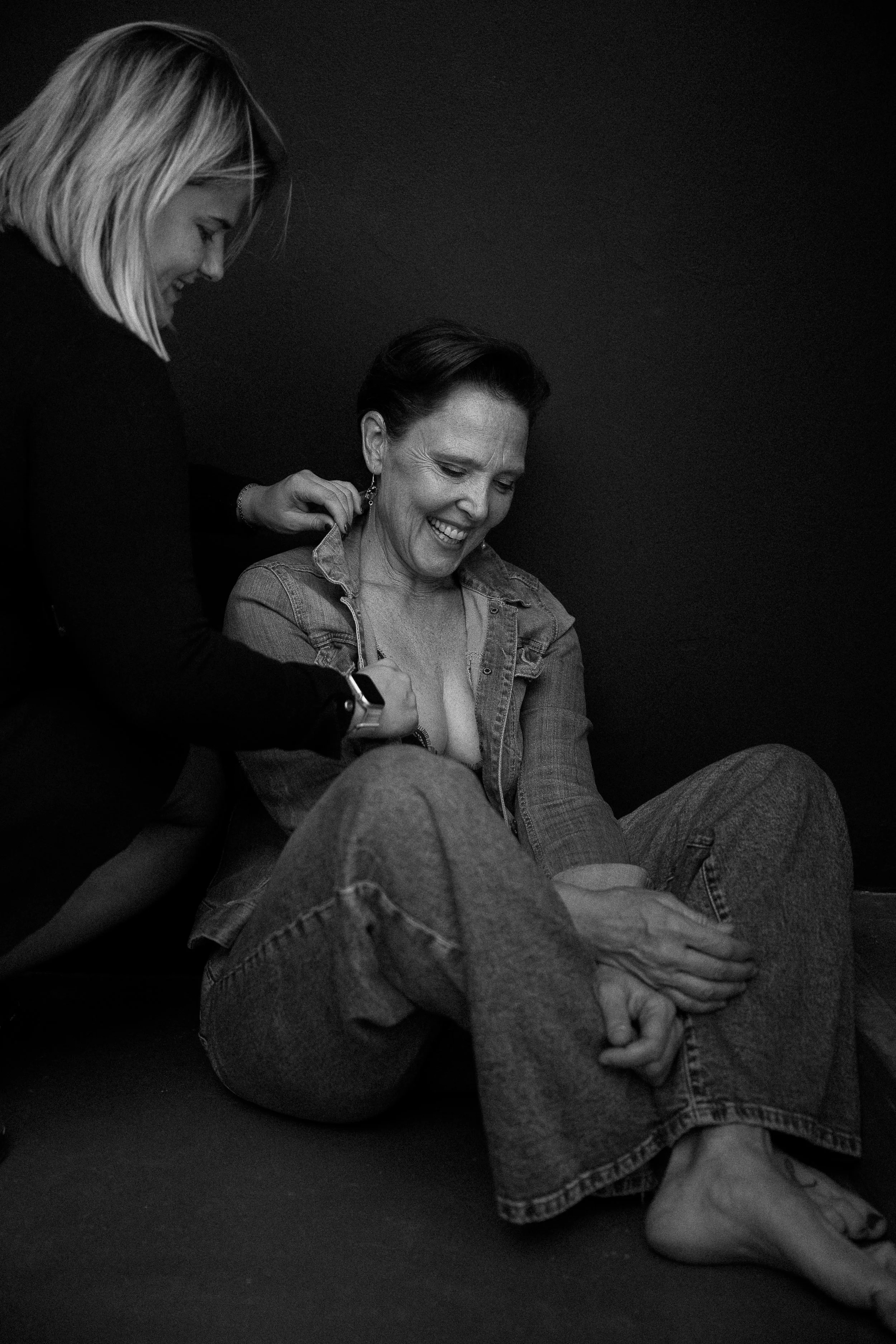 Twee vrouwen samen: één zit op de vloer, de andere staat achter haar en raakt haar oorbel aan — een warm, zorgend en zacht portretmoment.